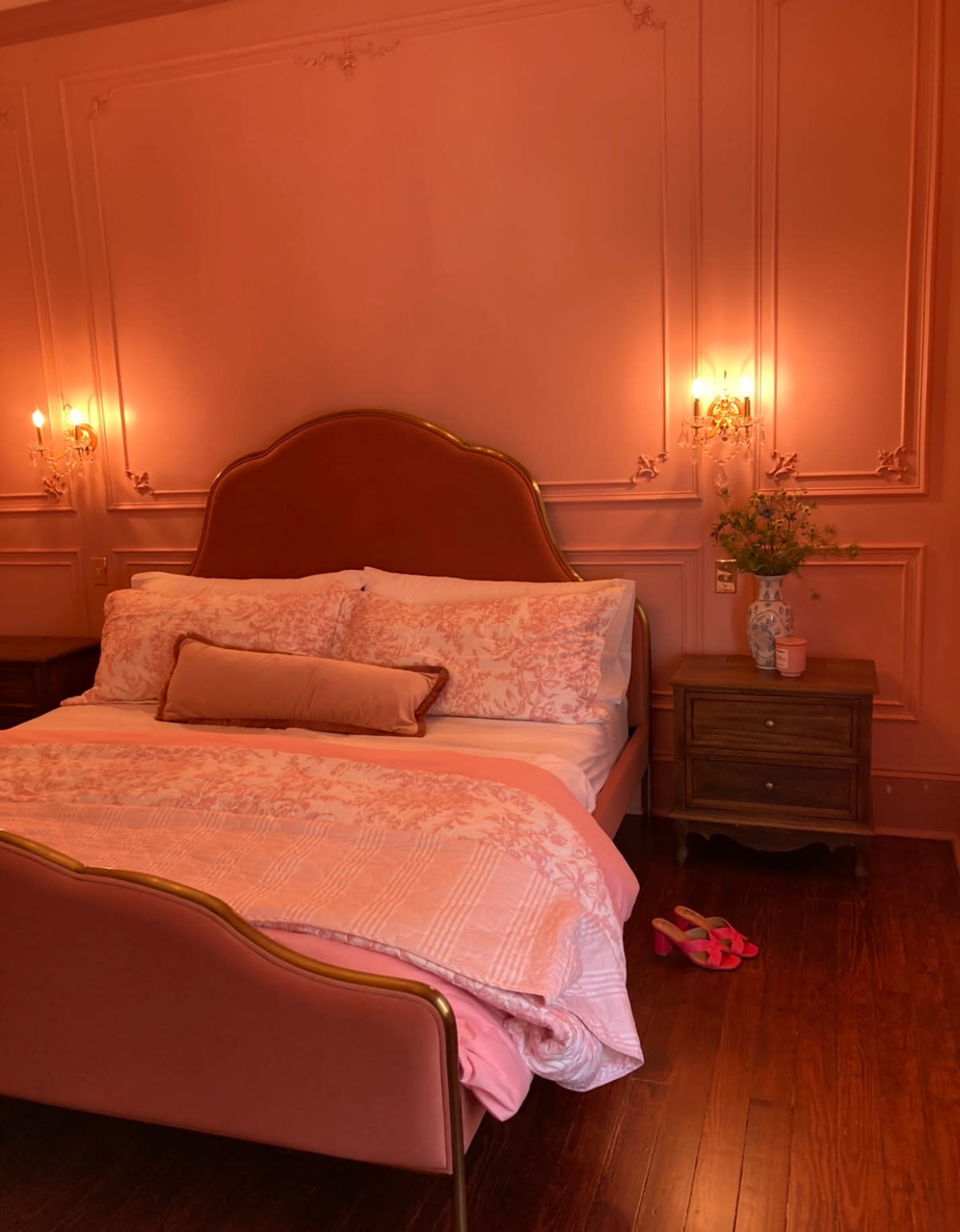 The image shows a softly lit bedroom featuring a pink-themed decor with a large bed, a bedside table, and a pair of pink slippers on the floor.