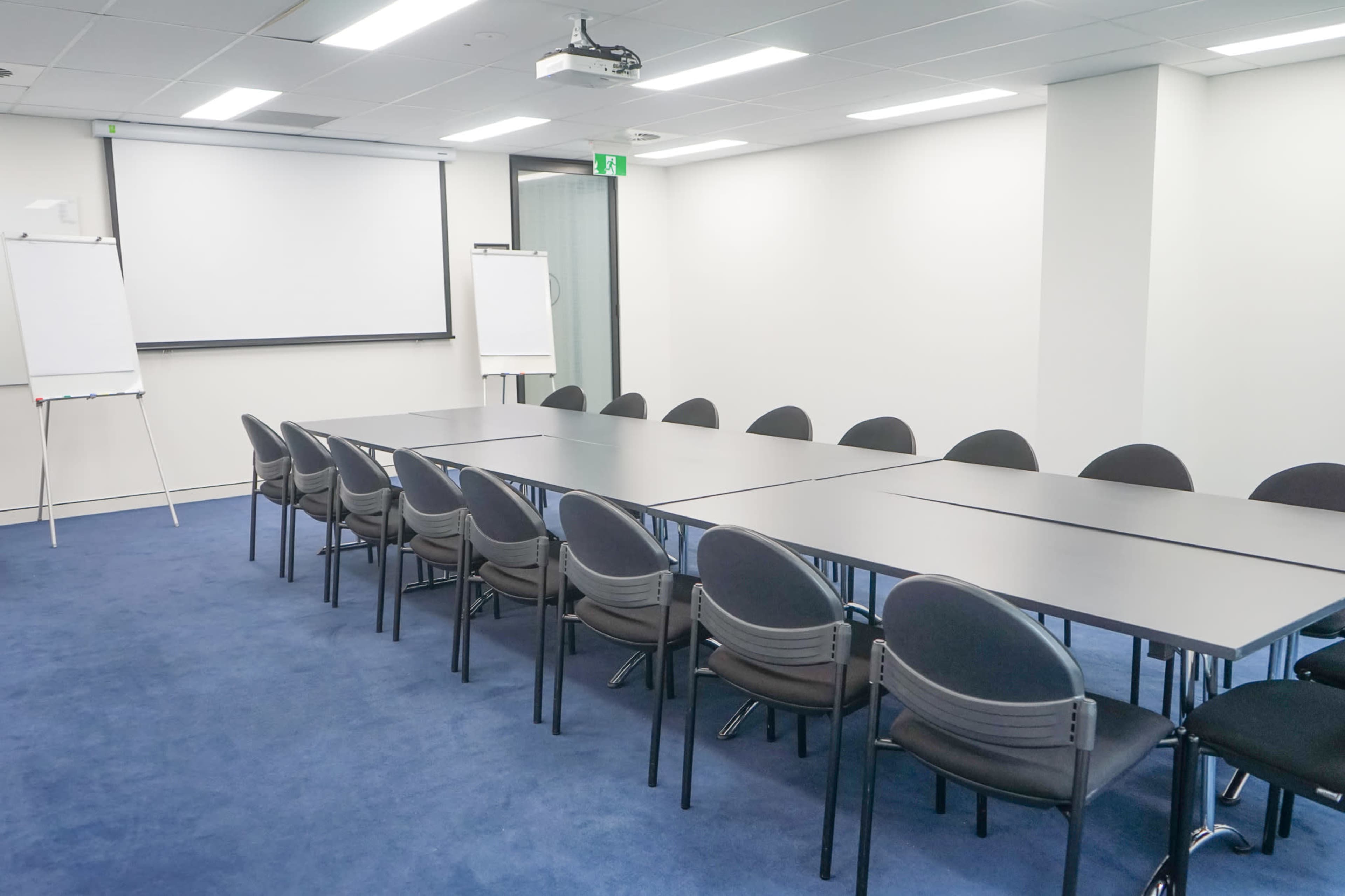 A conference room features a long table surrounded by black chairs, with a projector screen and whiteboards in the background.