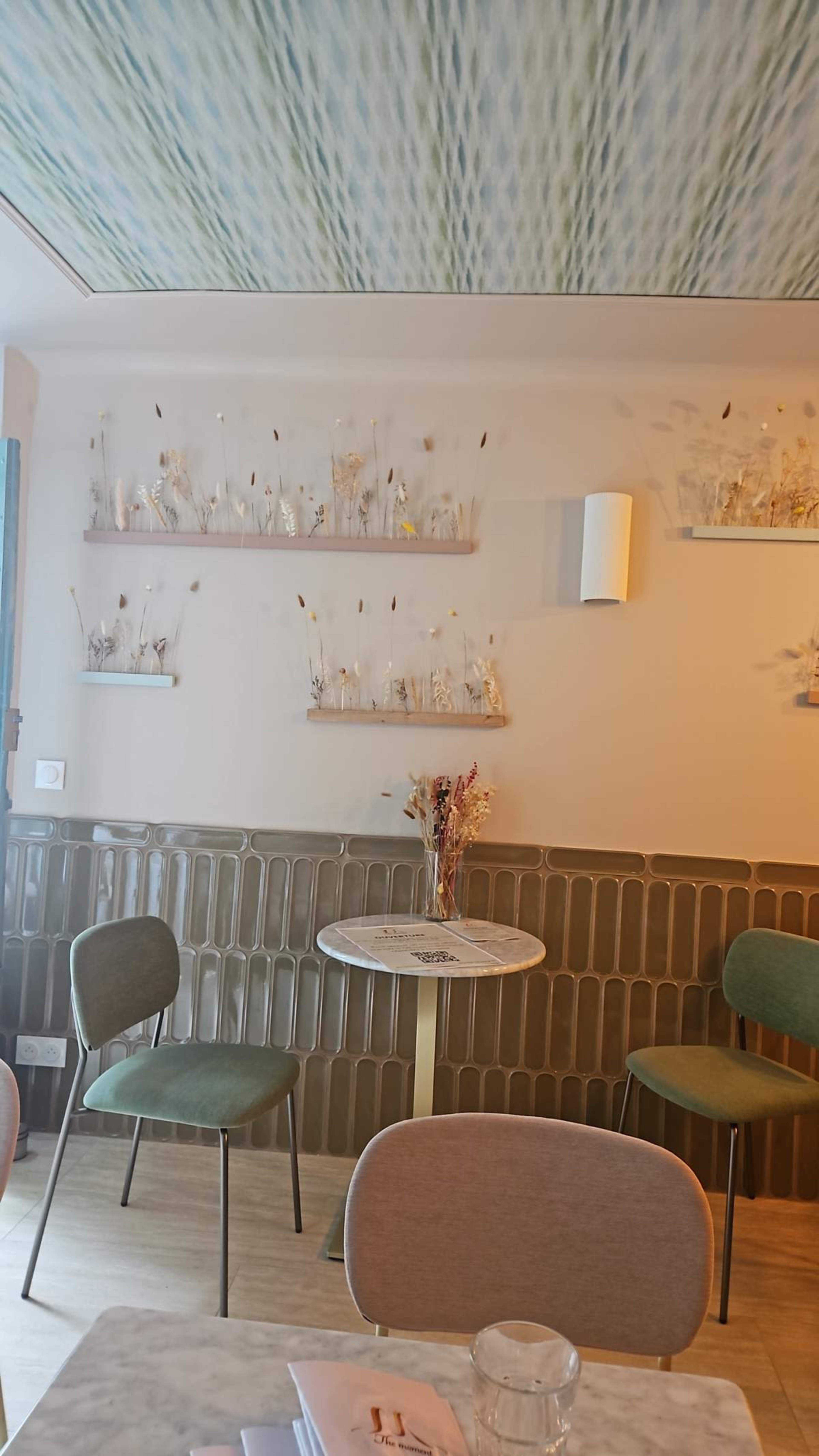 The image shows a cozy café interior with a marble-top table, two green chairs, and decorative shelves with dried flowers along the wall.
