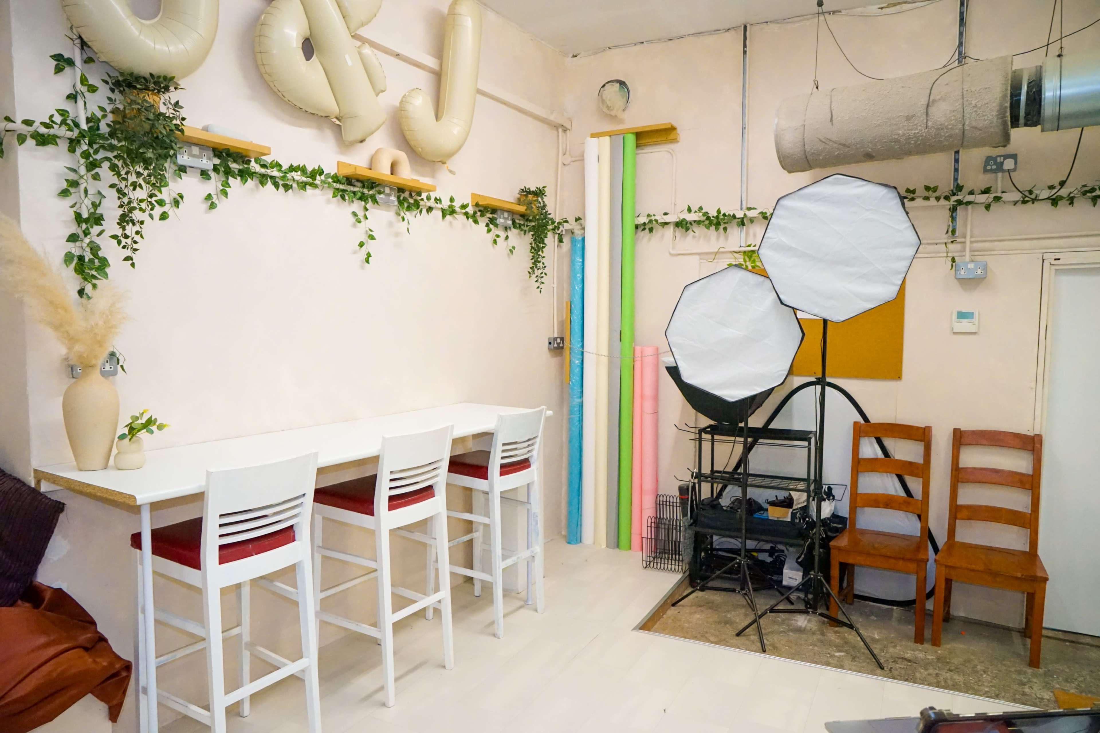 The image shows a studio space with a white bar table and stools, decorated with plants, and featuring professional lighting equipment and colorful backdrops against a light-colored wall.