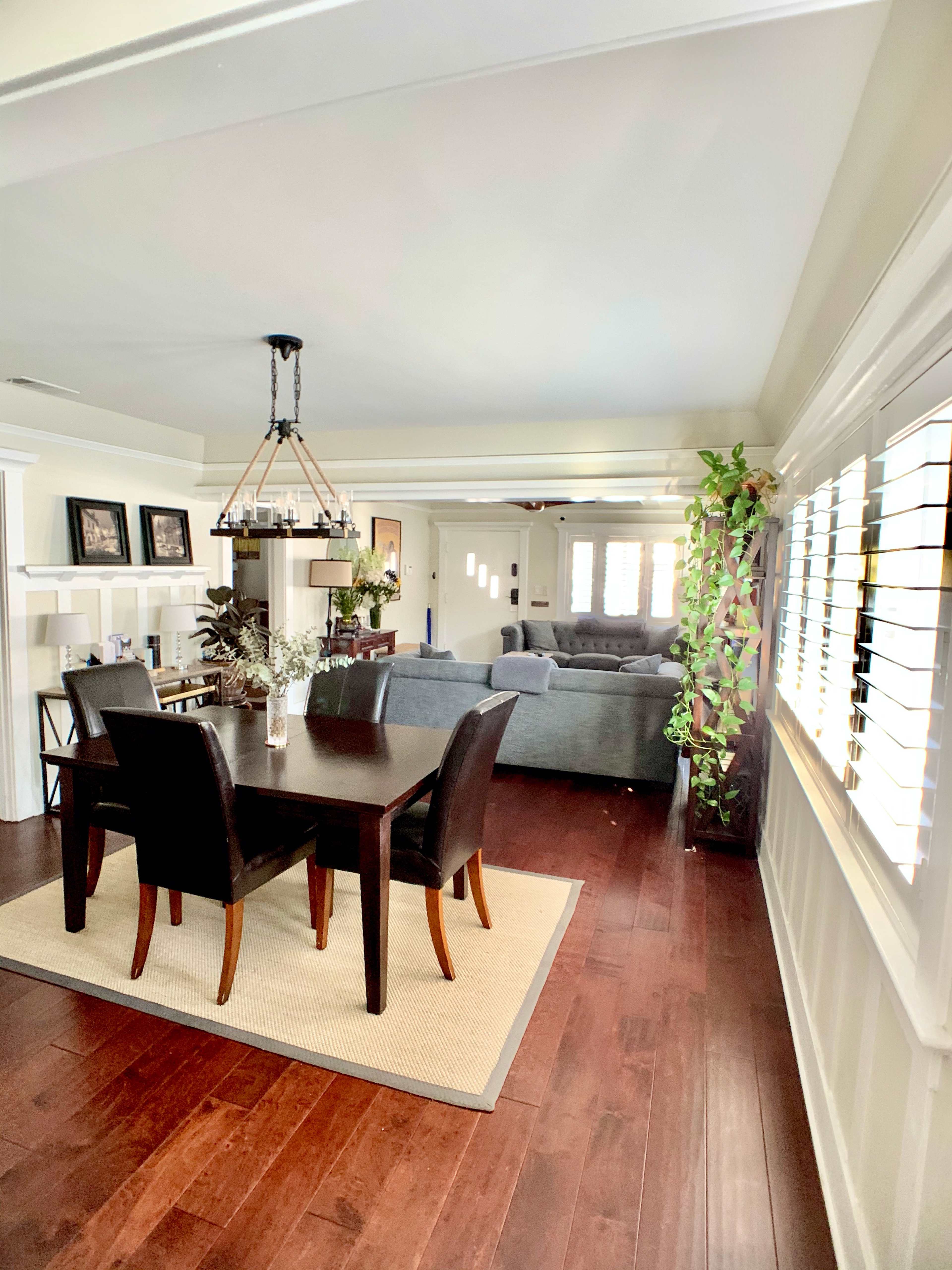A living space with a dining area featuring a dark wooden table and black chairs, leading into a living room with a gray sofa and multiple windows.