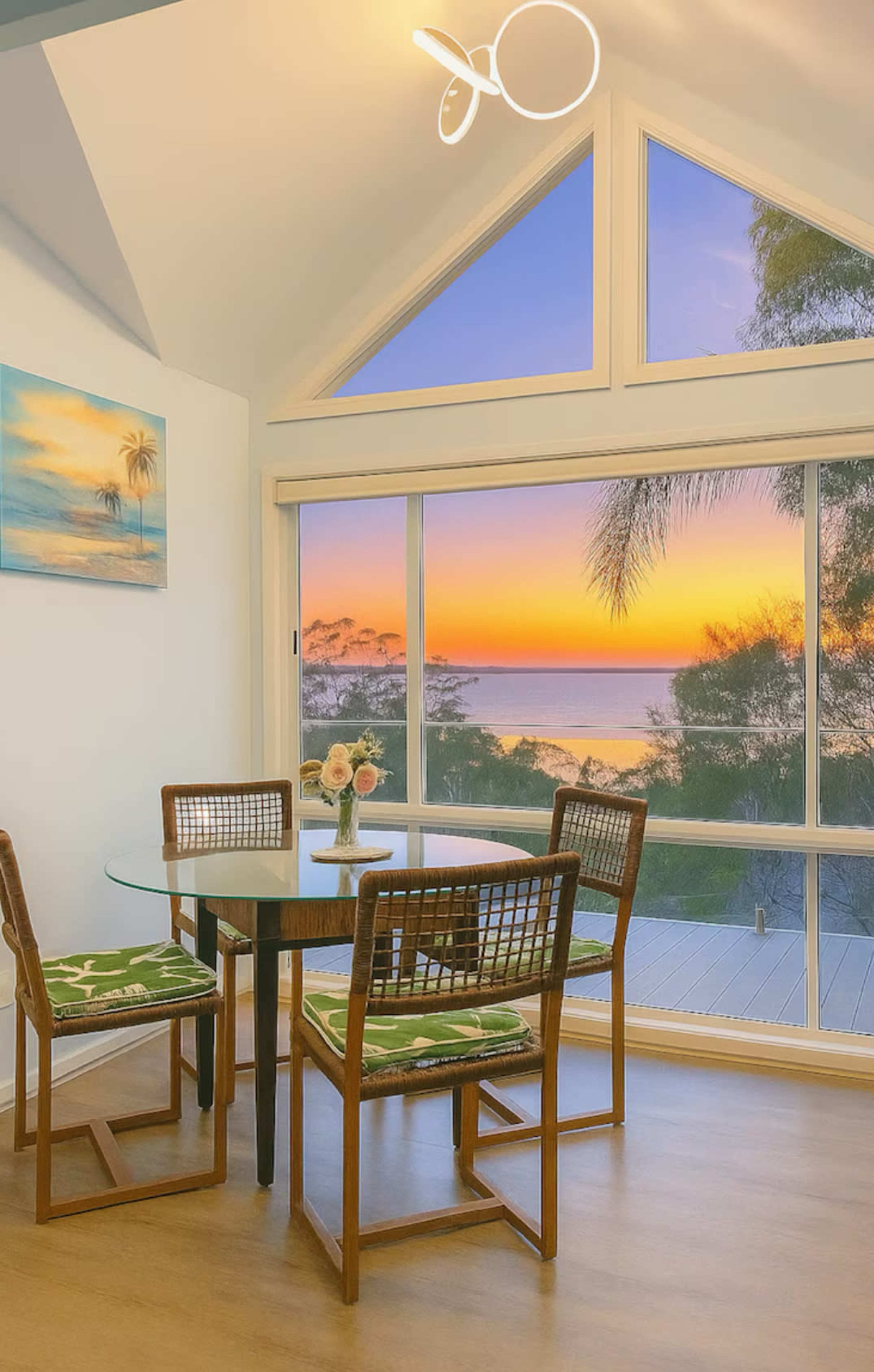 A dining area features a glass table with wooden chairs and a view of a sunset over the ocean through large windows.