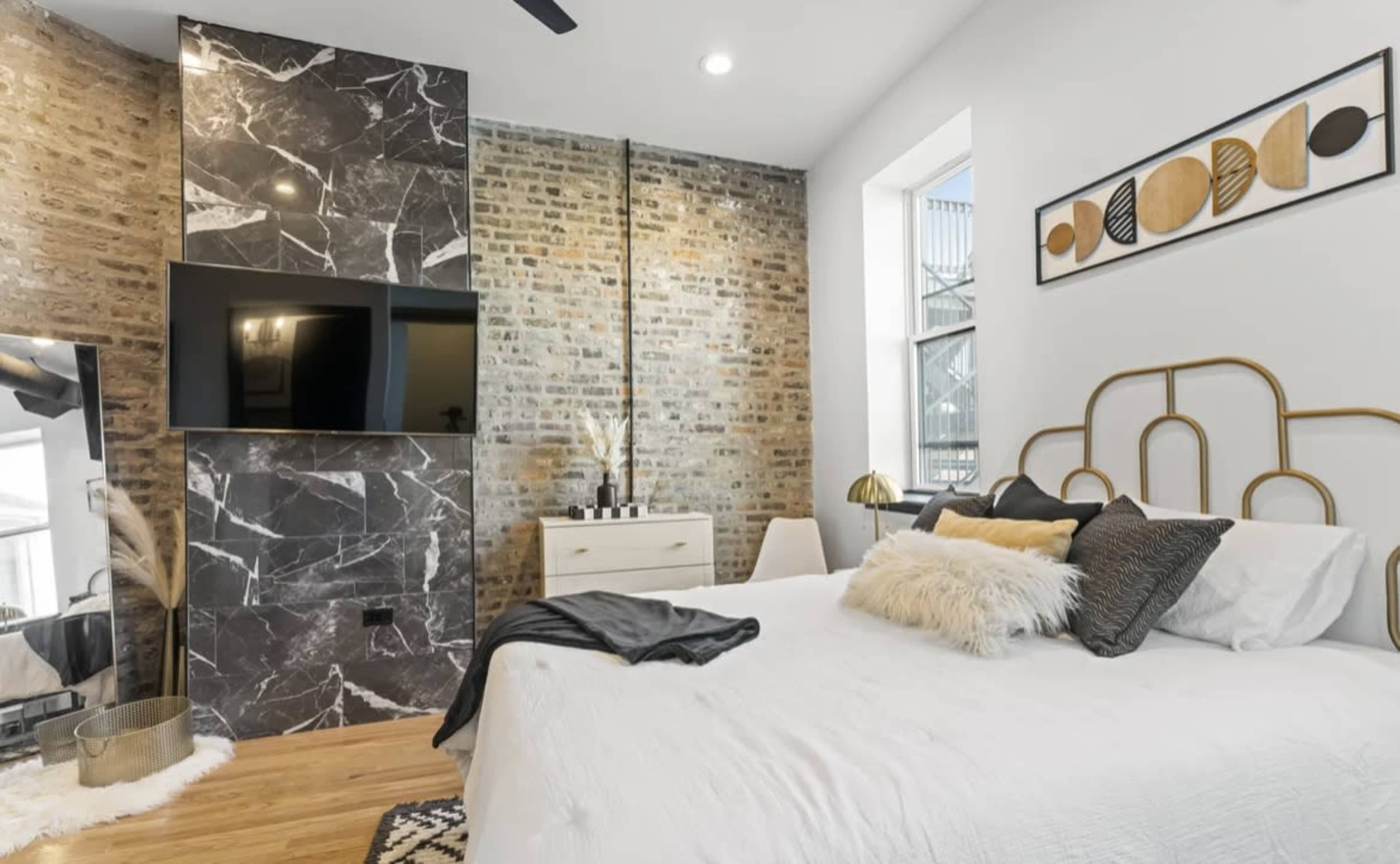 A modern bedroom with a white bed, a television mounted on a marbled wall, and exposed brick accents.