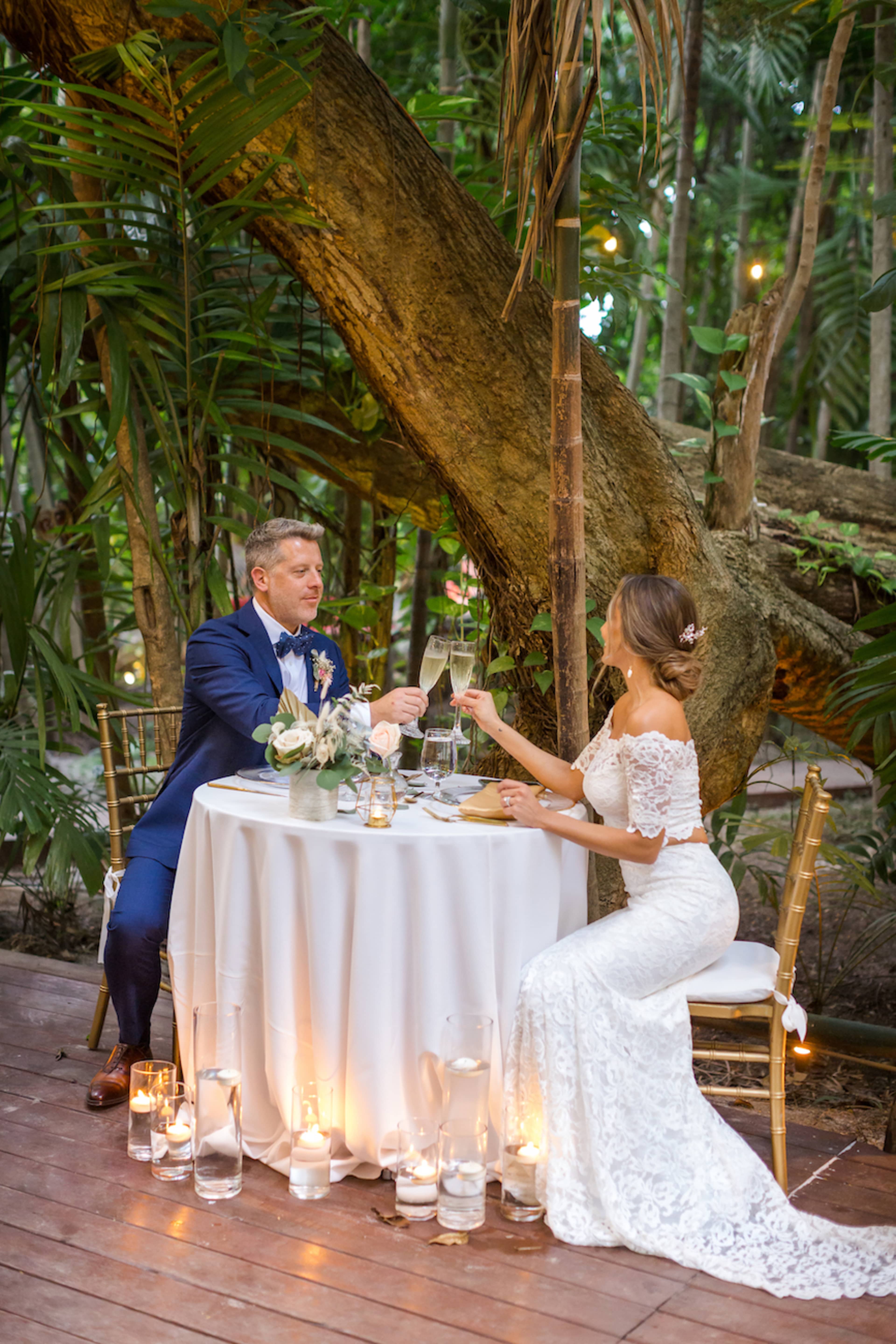 A couple toasts at an elegantly set table beneath a large tree in a lush, green outdoor setting.