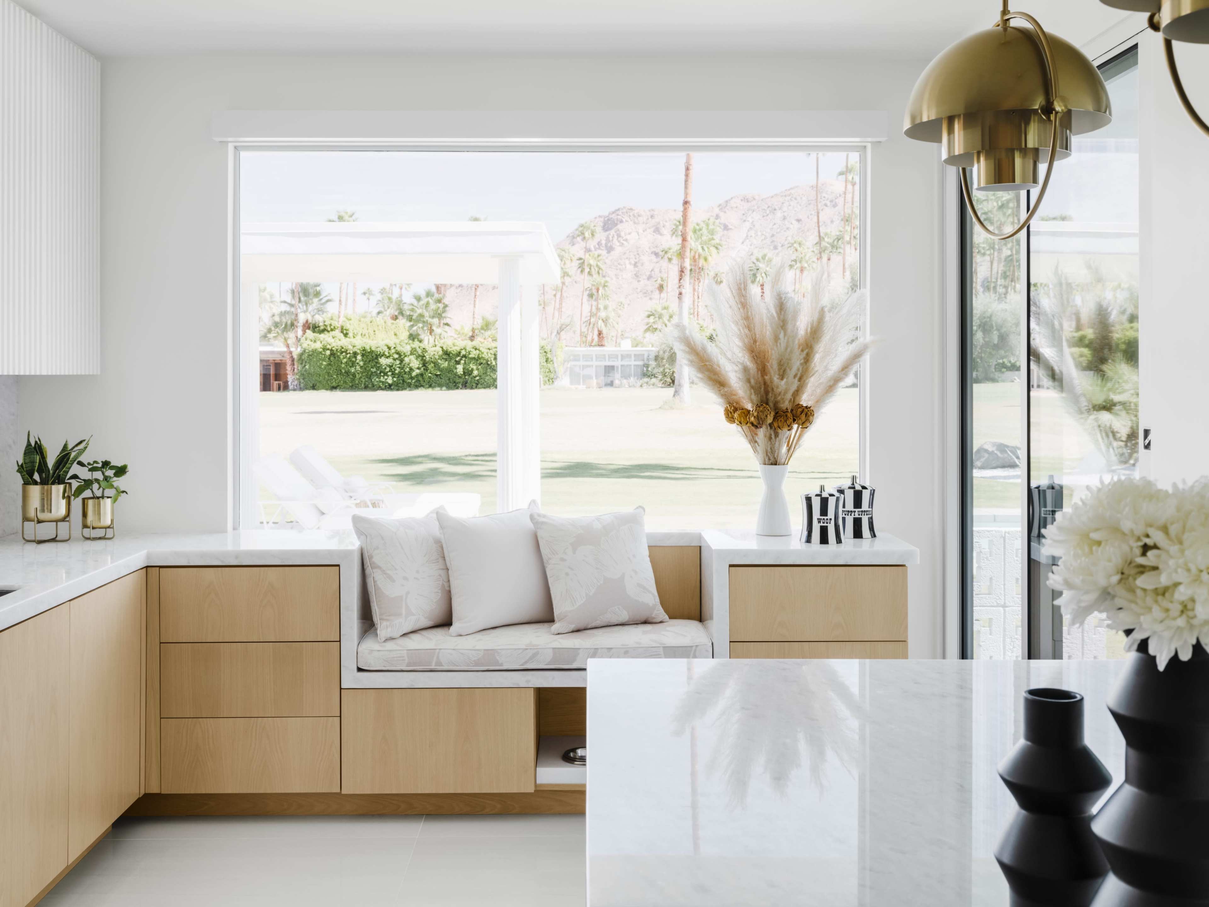 A modern kitchen features a window seat with pillows, overlooking a landscaped outdoor area with palm trees and a view of mountains in the background.