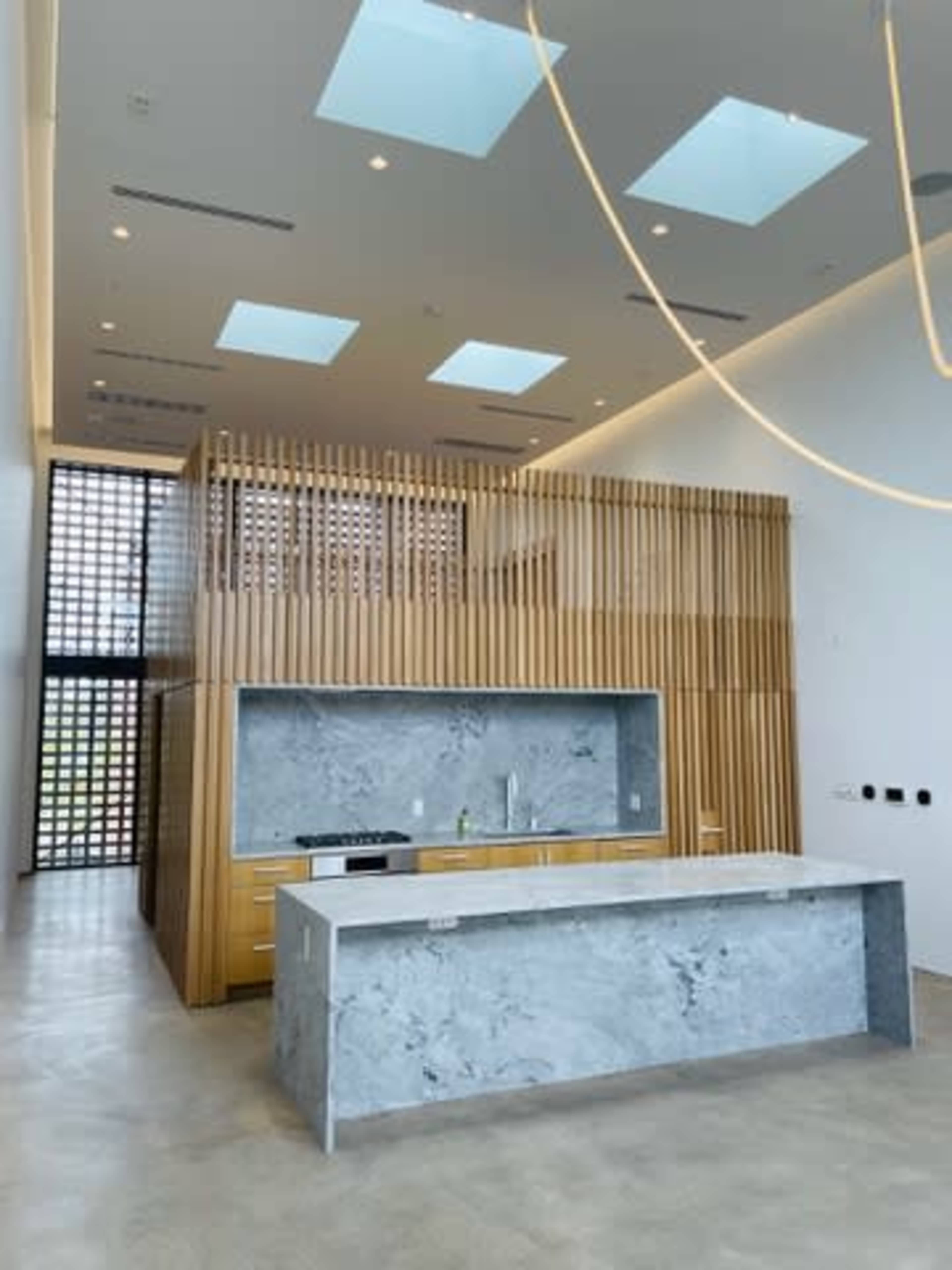 The image shows a modern kitchen with a large marble island and a wooden partition behind it, featuring several skylights above.