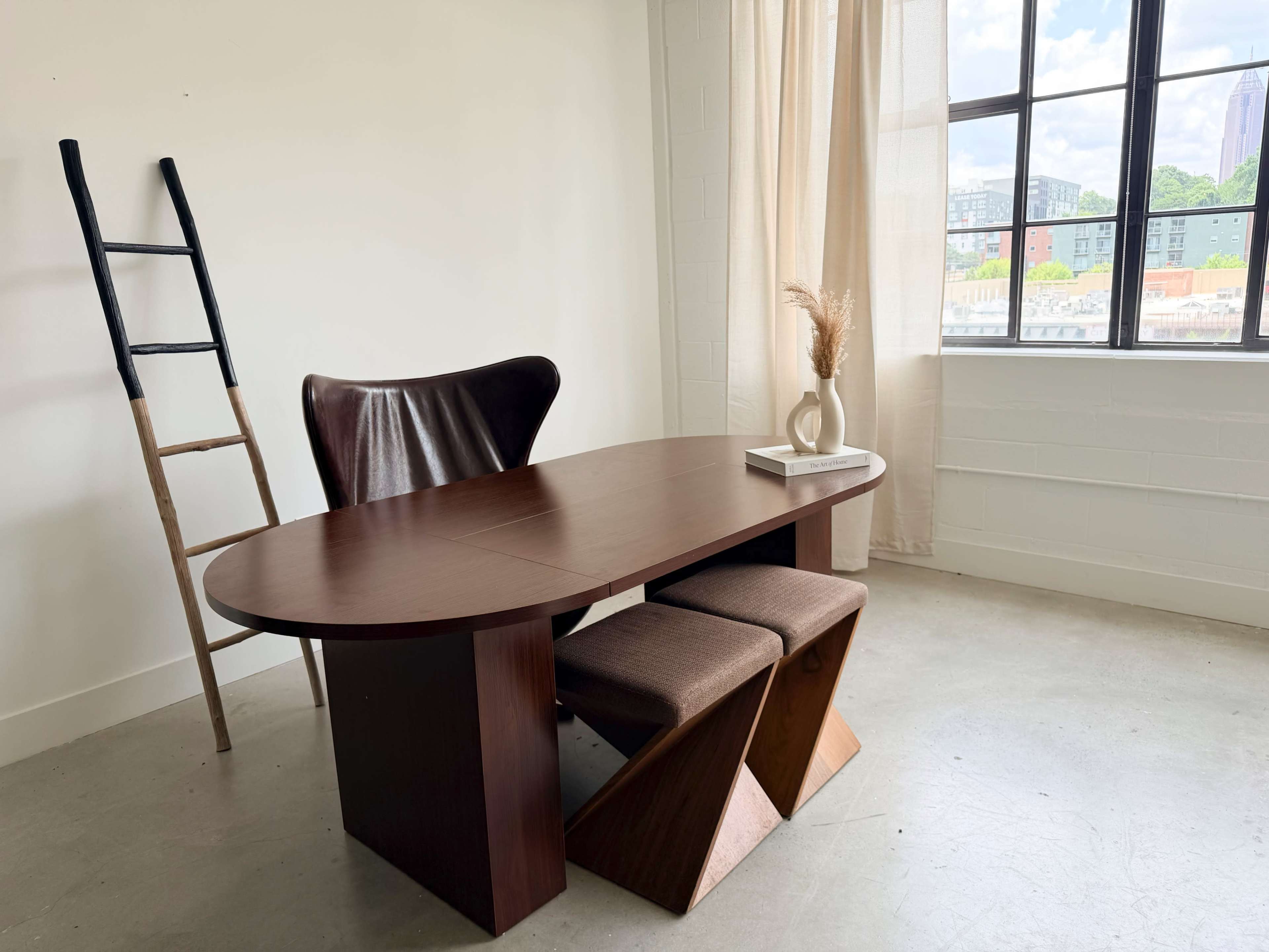 The image shows a modern dining area featuring a wooden table with two stools and a decorative vase near a large window.