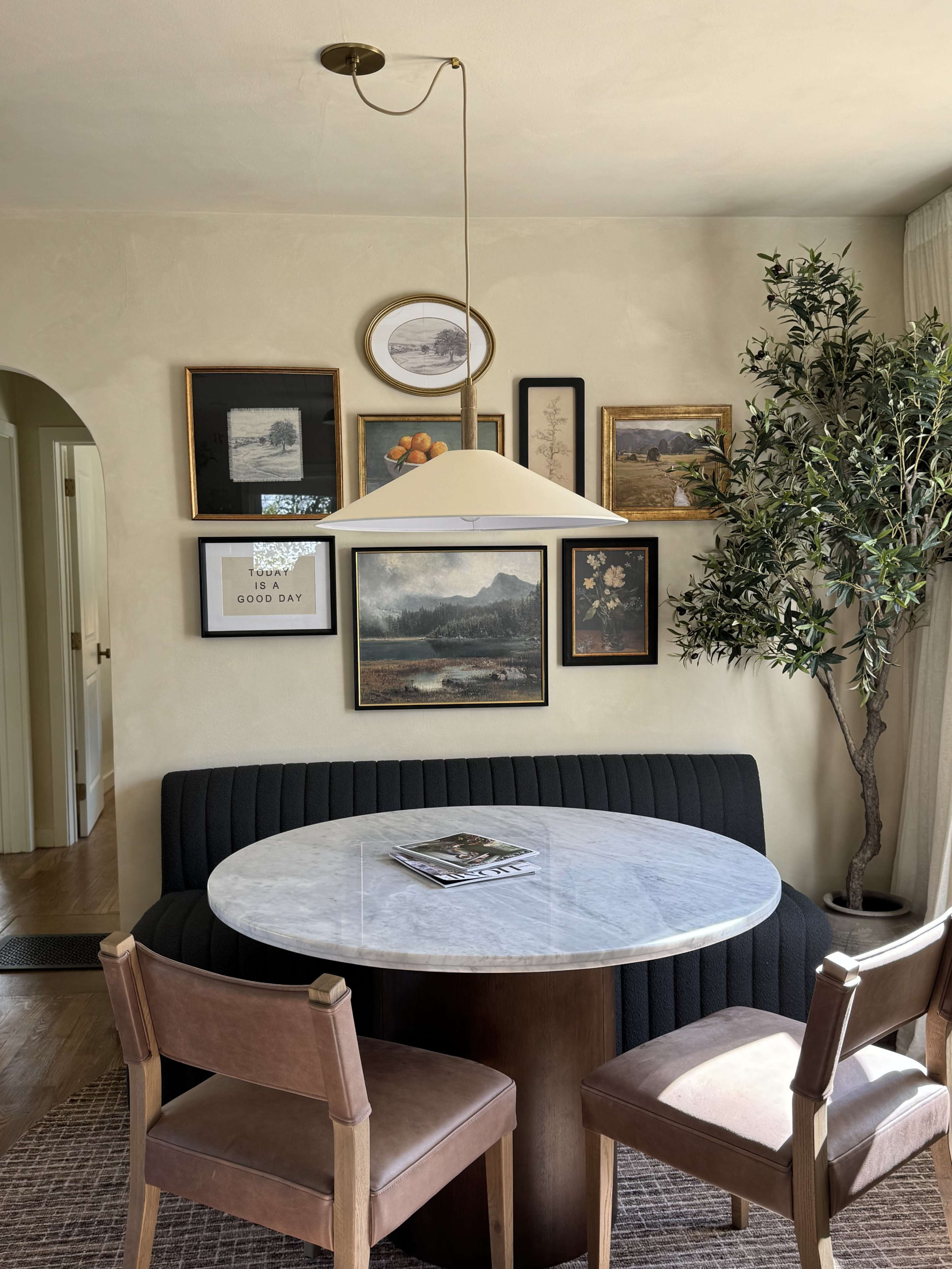 The image shows a dining area with a round marble table, two brown chairs, and a gallery wall featuring various framed artworks.