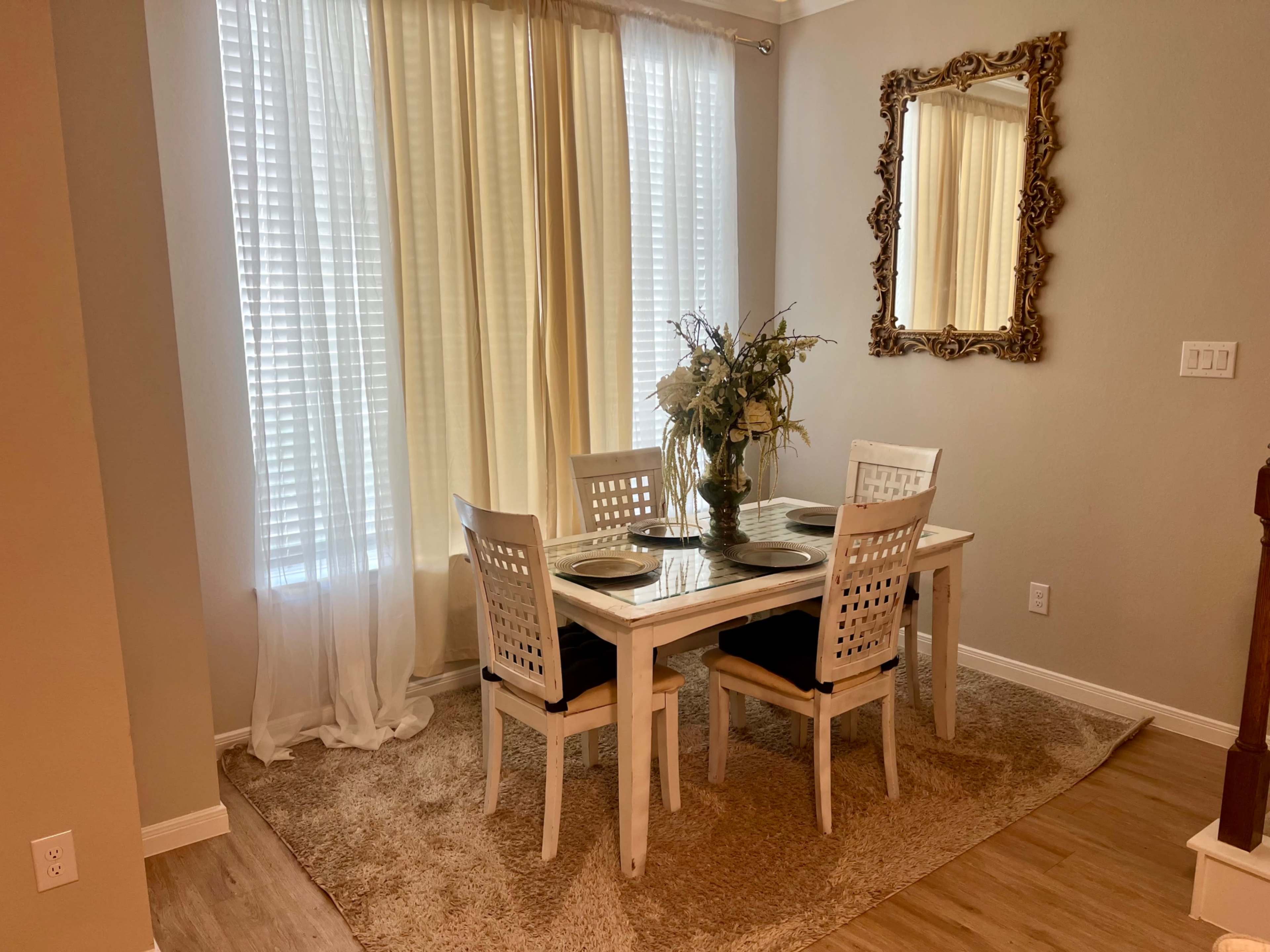 A dining table with four chairs is arranged in a well-lit room, featuring large windows draped with light curtains and a decorative mirror on the wall.