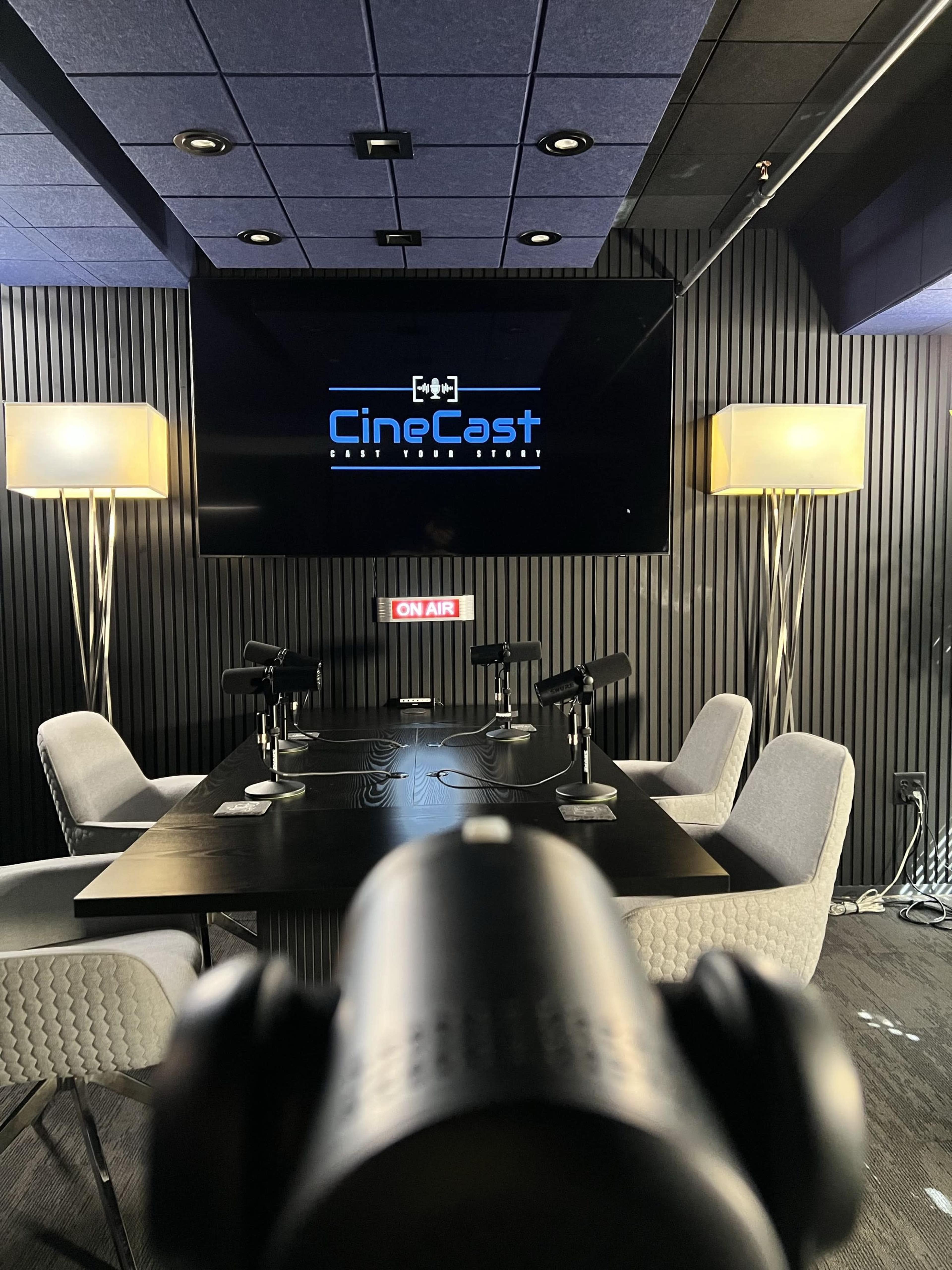 A podcasting studio features a large screen displaying the "CineCast" logo, multiple microphones on a dark wooden table, and modern chairs under ambient lighting.