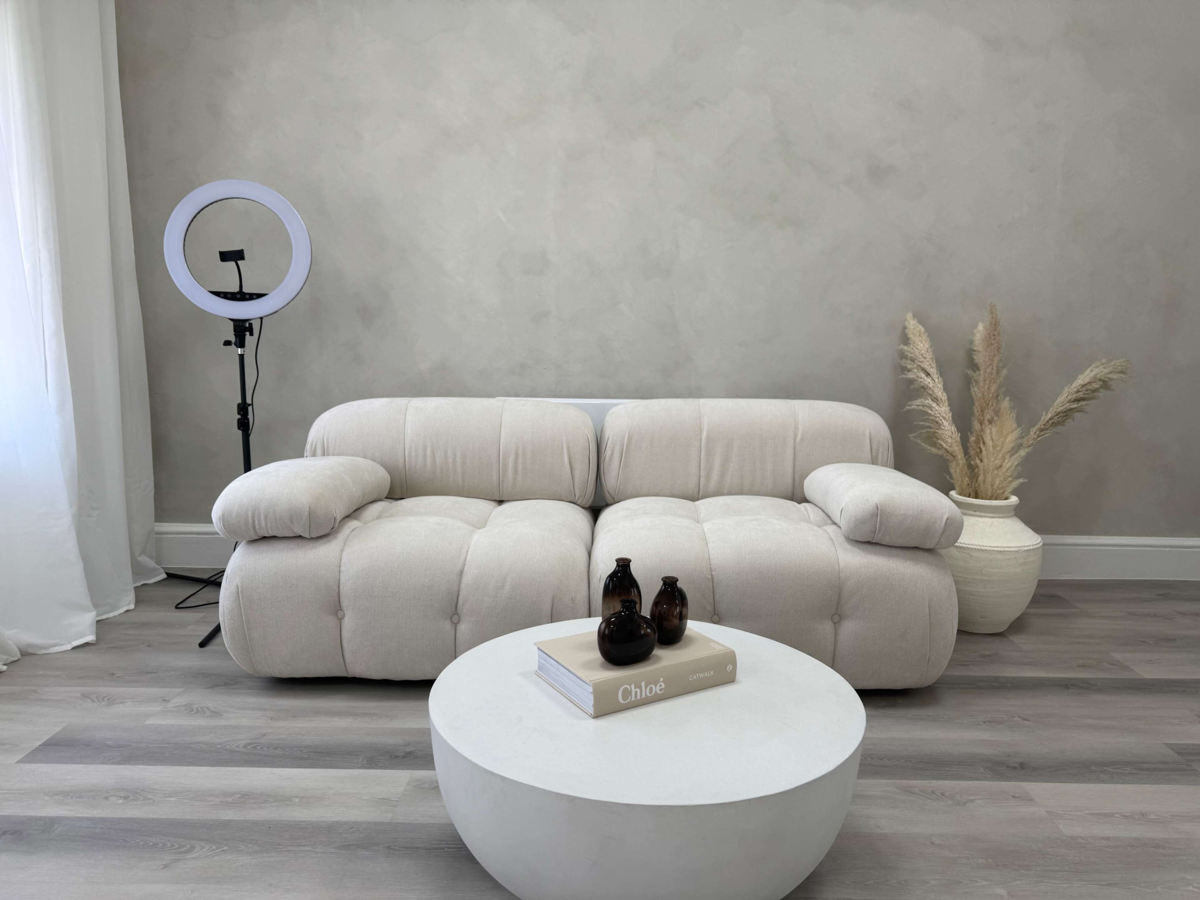 A light-colored, modern sofa is positioned in a minimalist room with a round white coffee table and decorative items, along with a ring light in the background.