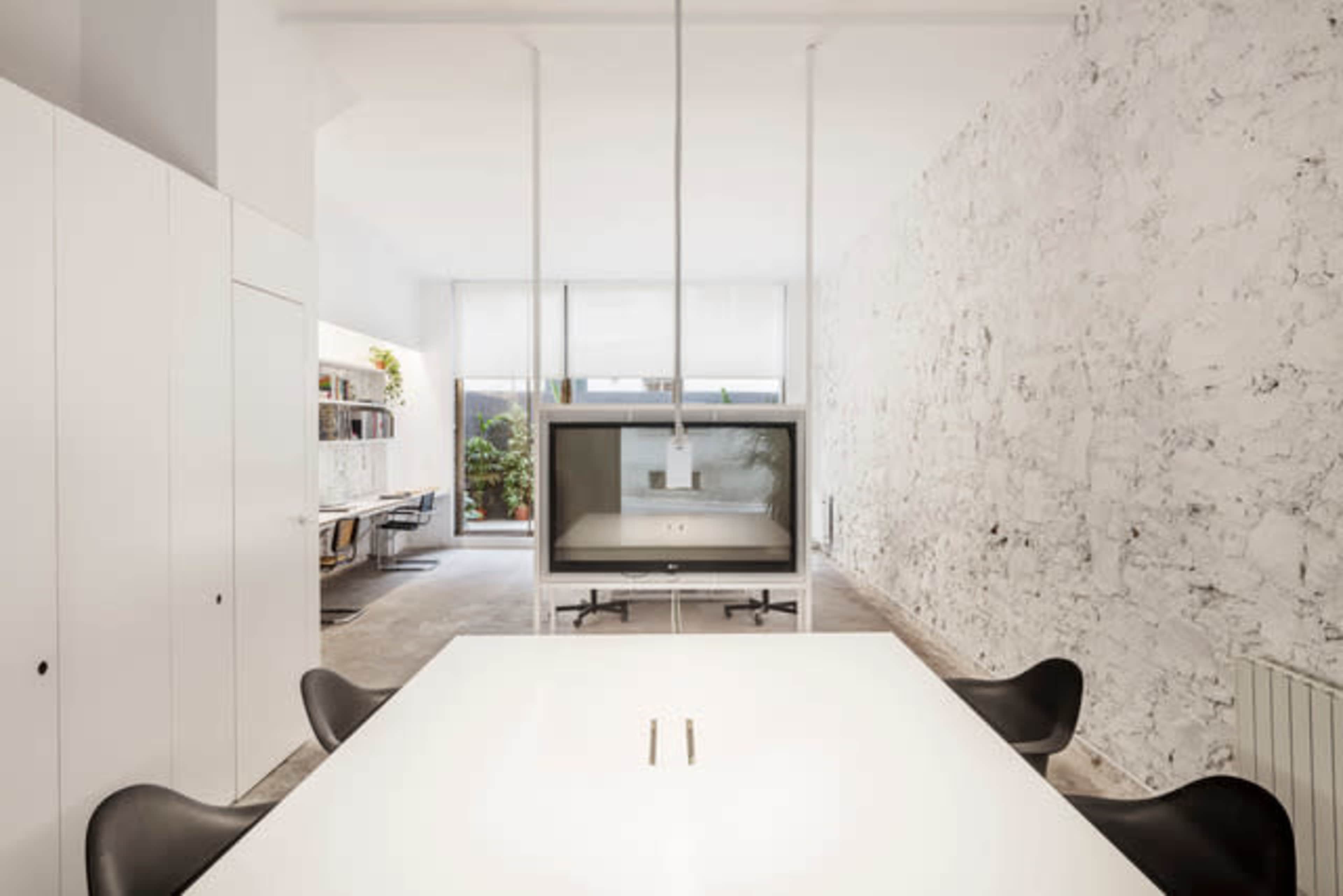 A modern, minimalist office space with a large white table and a view of a workstation area featuring glass walls and greenery.