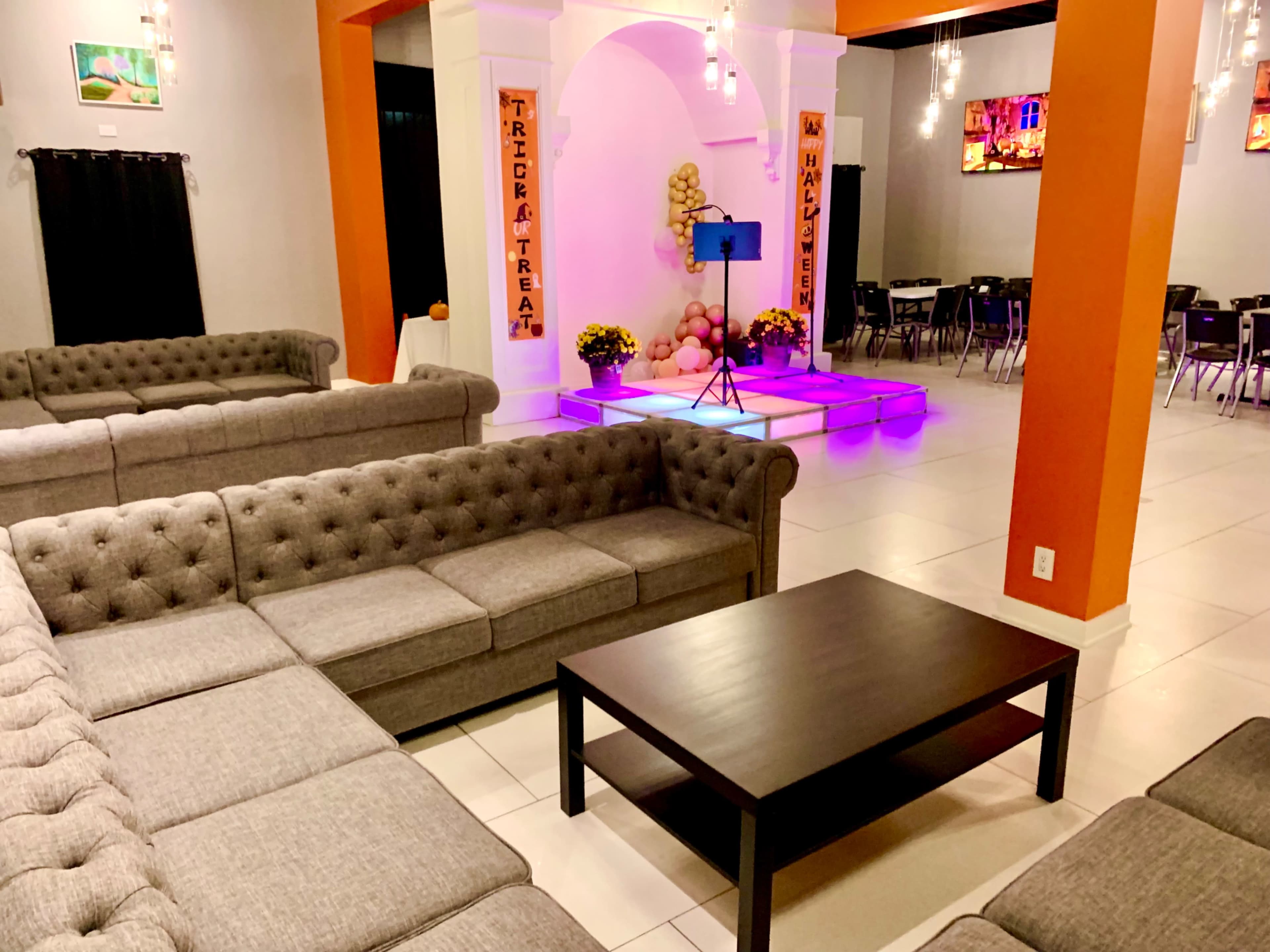 The image shows a modern event space featuring gray tufted couches arranged around a black coffee table, with a stage area illuminated in purple light and decorated with colorful artwork.