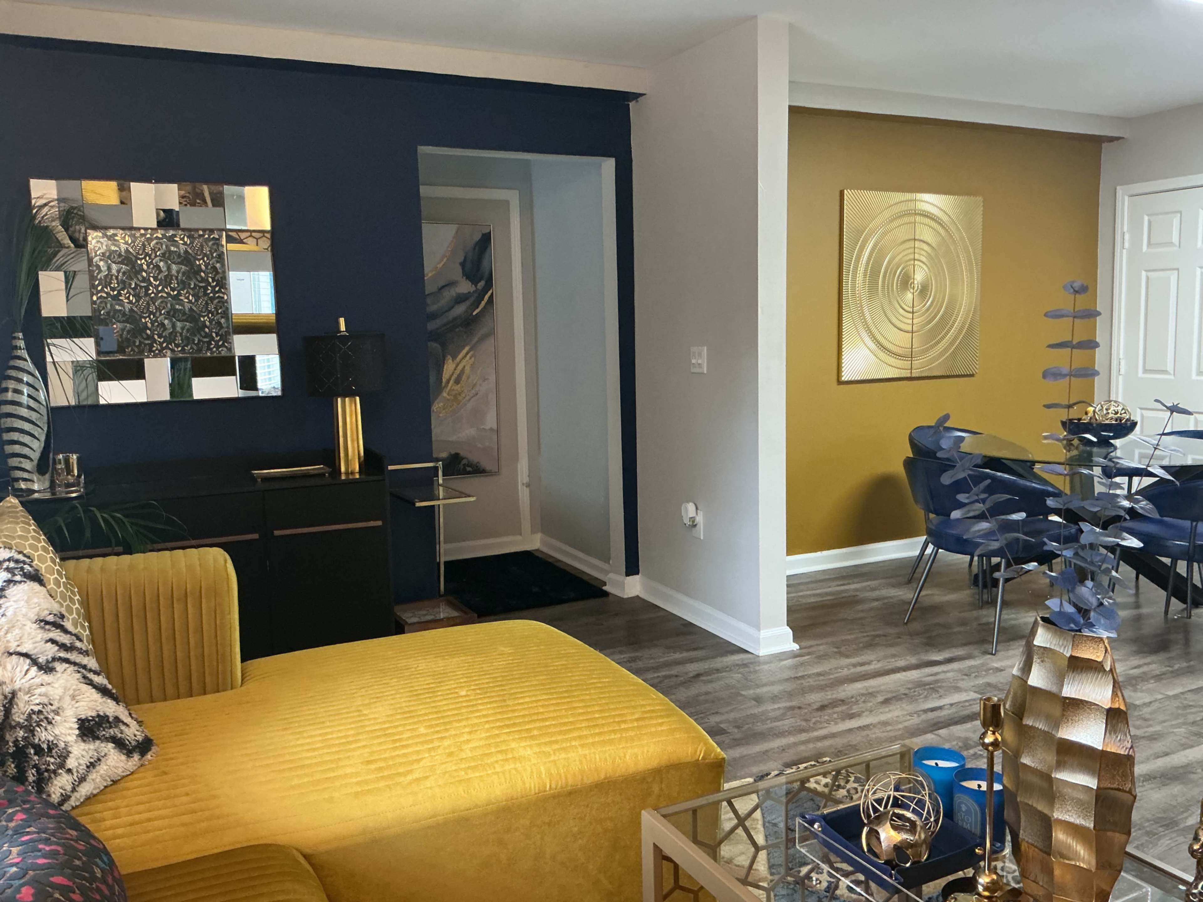 The image shows a modern living space featuring a yellow sofa, an abstract wall art piece, and a dining area with a round table and blue chairs.