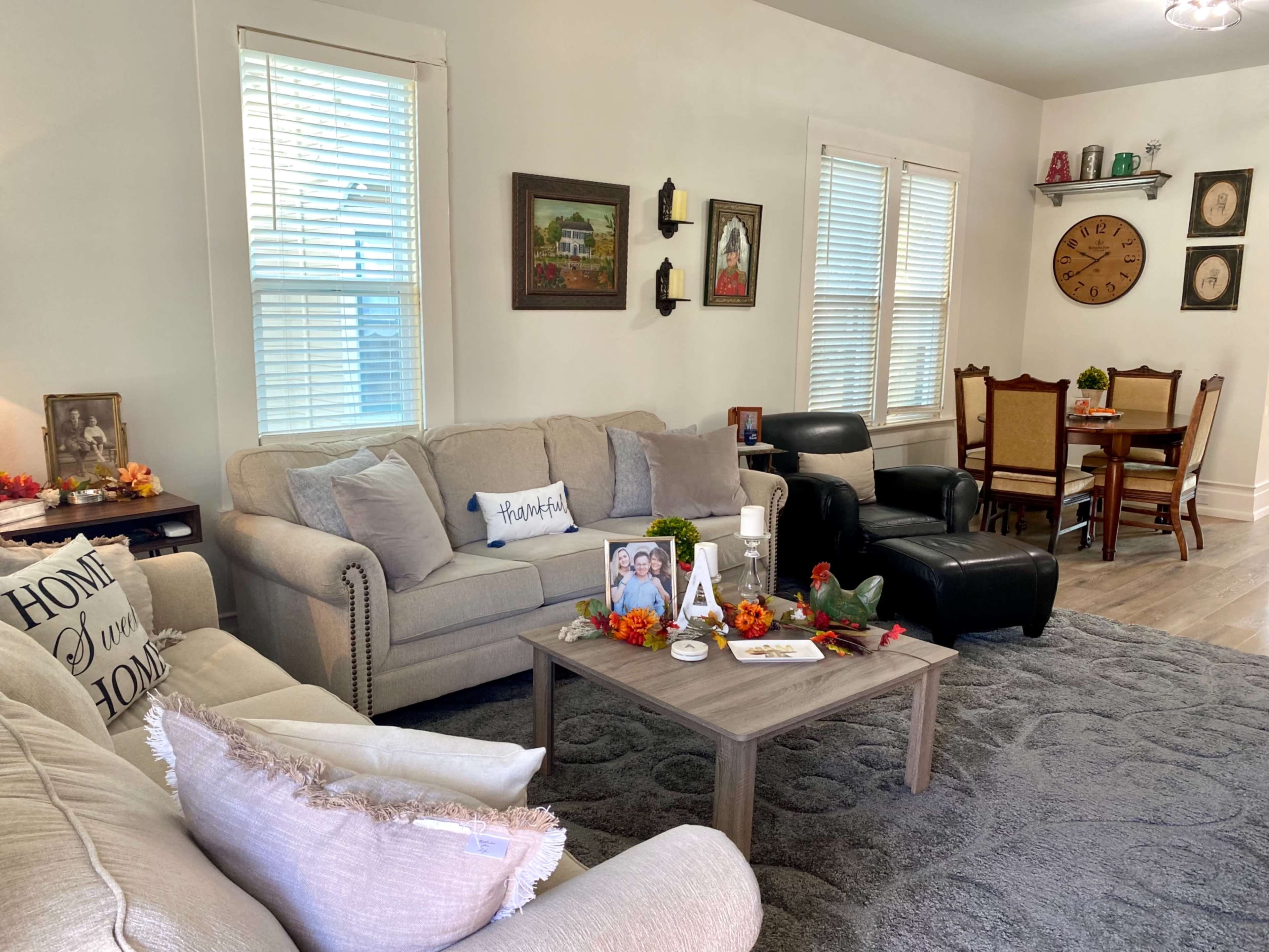 A cozy living room features a beige sofa, a dark armchair, a coffee table adorned with decor, and a dining area in the background with a clock and seated chairs.