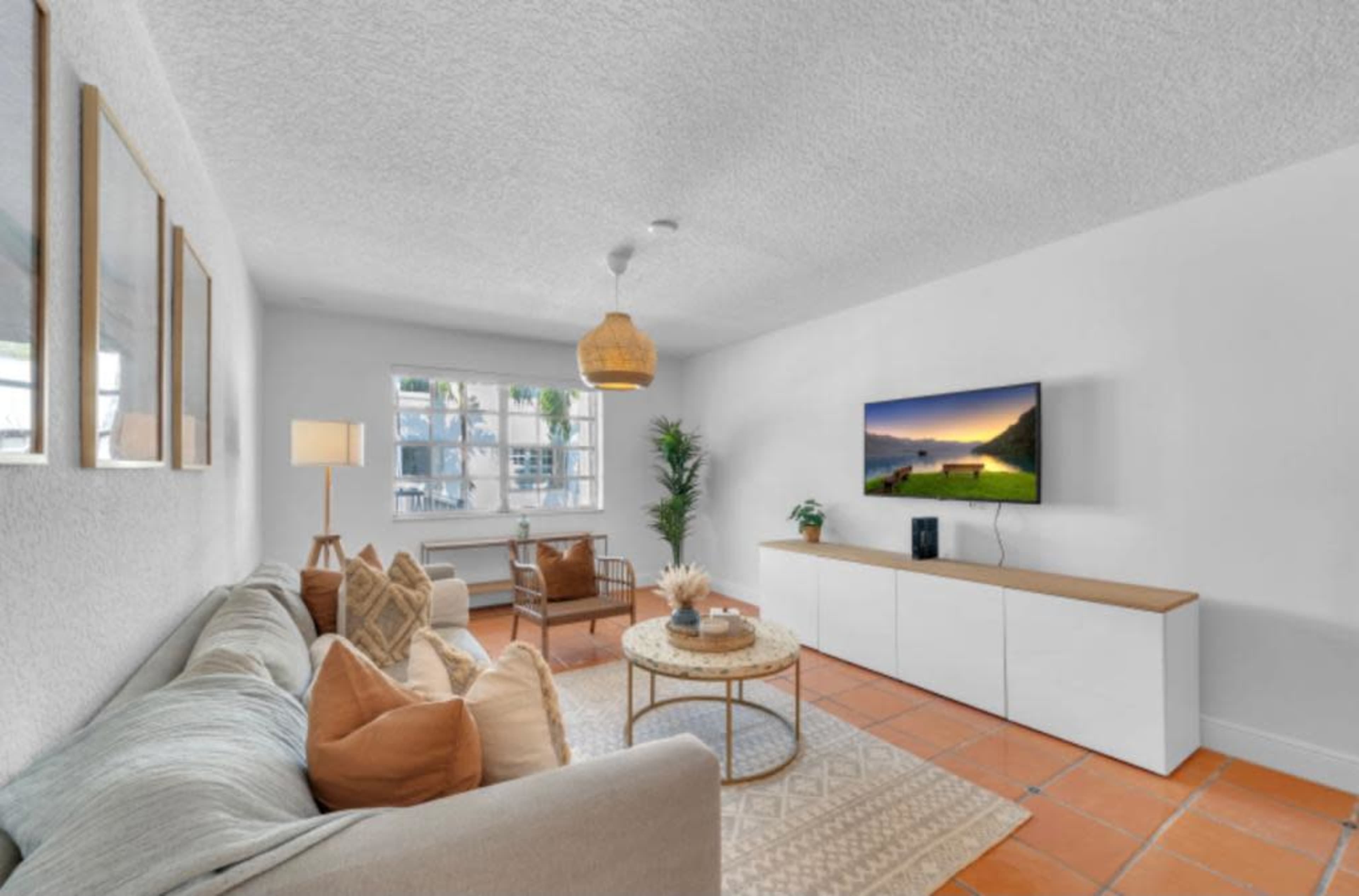 A living room features a sofa with decorative pillows, a coffee table, a television on a white console, and natural light from windows.