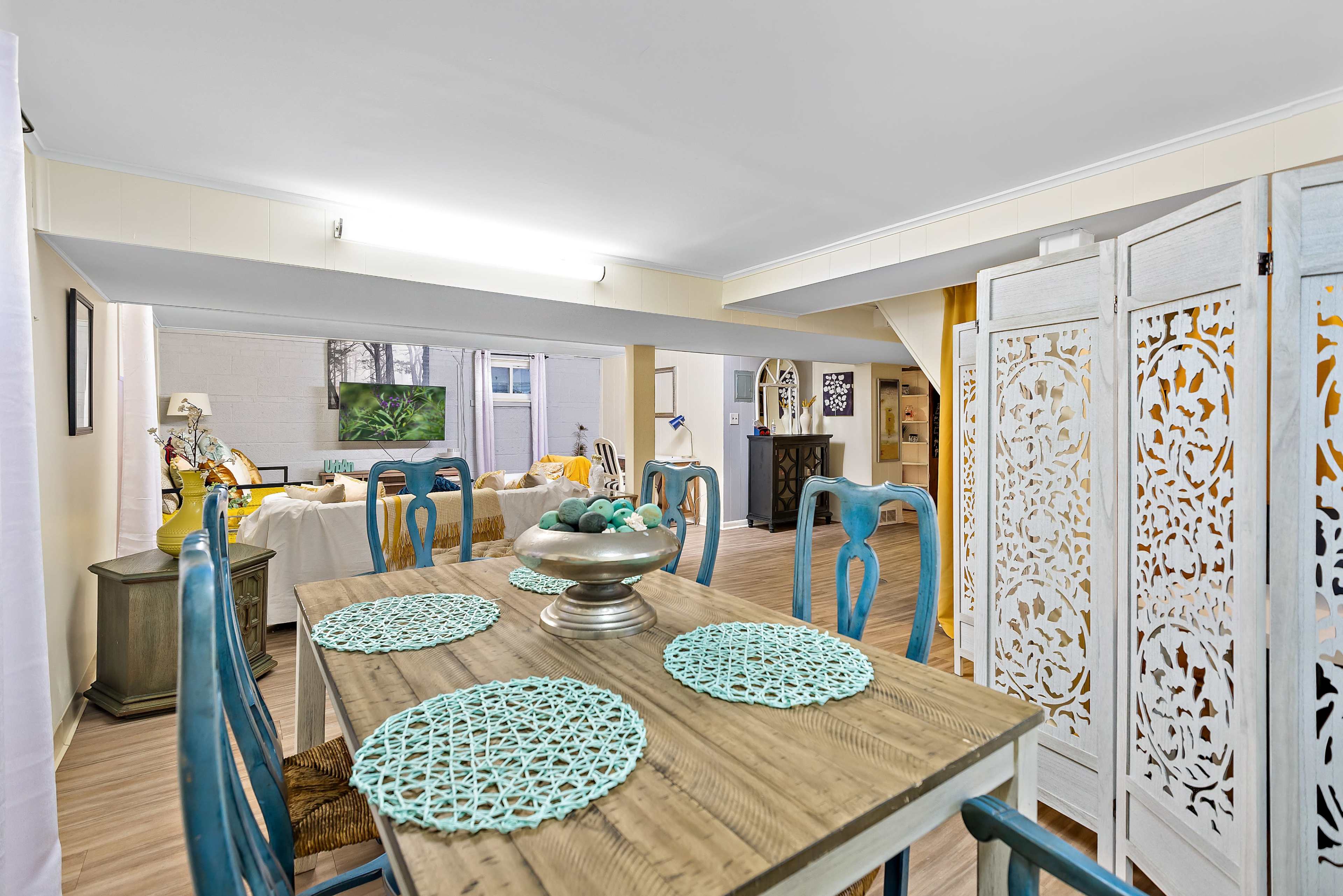 A dining area features a wooden table set with decorative placemats and a centerpiece, leading to an open living space in a softly lit interior.