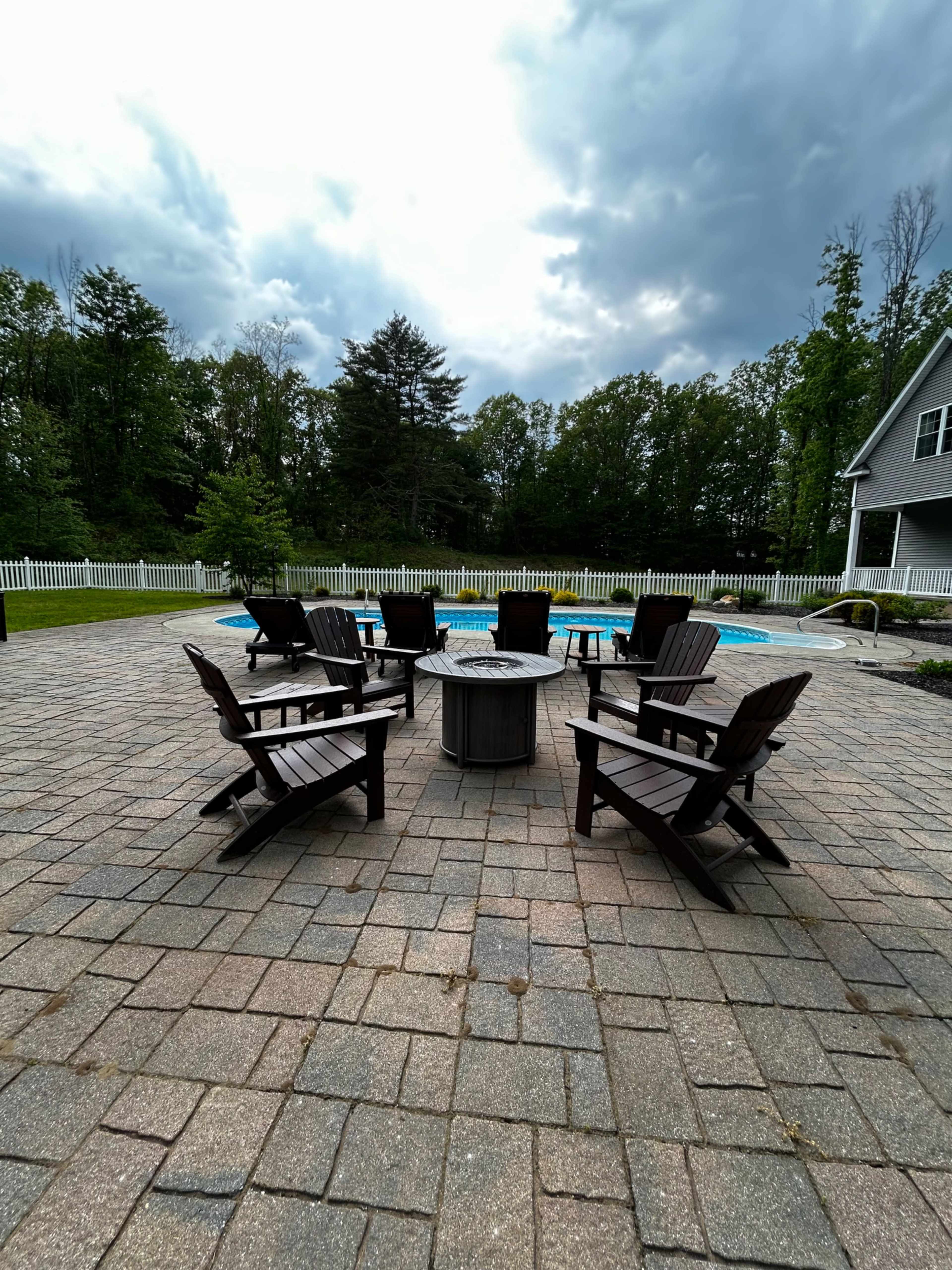A circle of wooden Adirondack chairs surrounds a fire pit on a patio near a swimming pool, with trees and a white fence in the background.