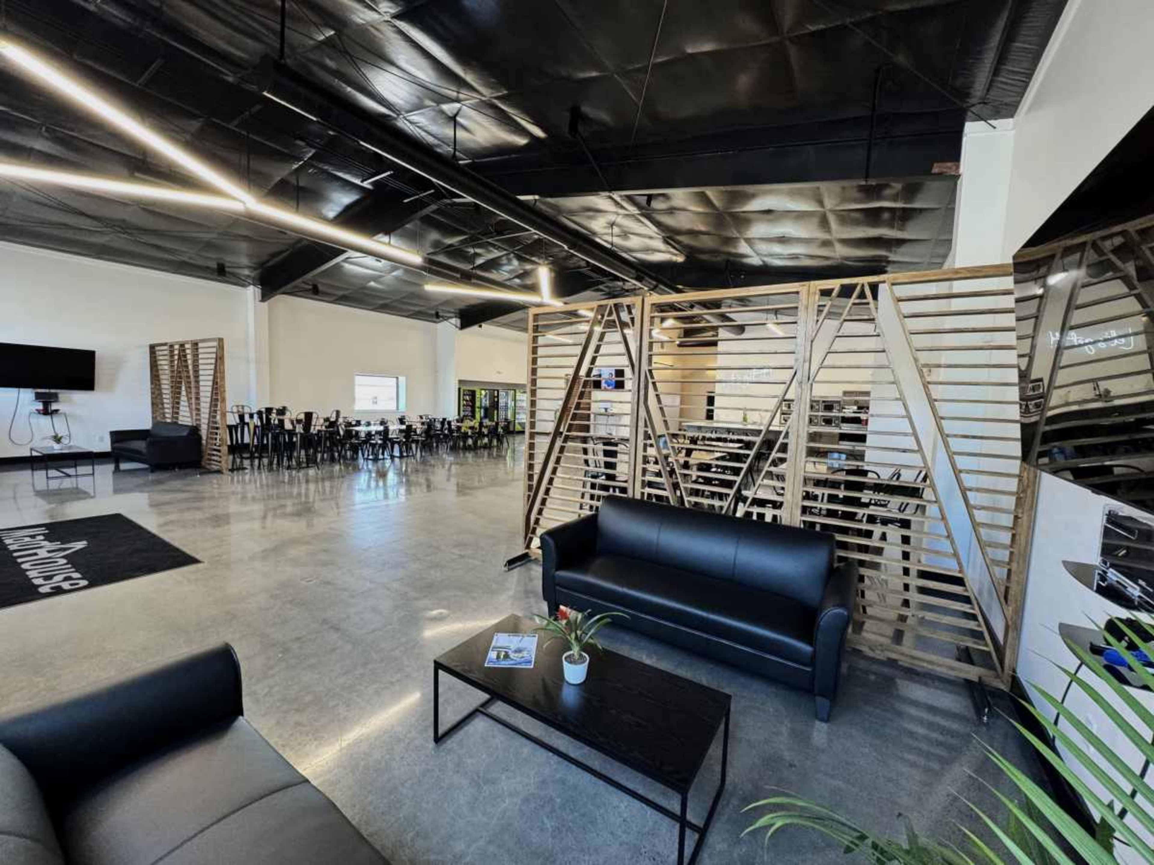 A modern open space with black leather couches, a wooden partition, and rows of tables and chairs in a well-lit environment.