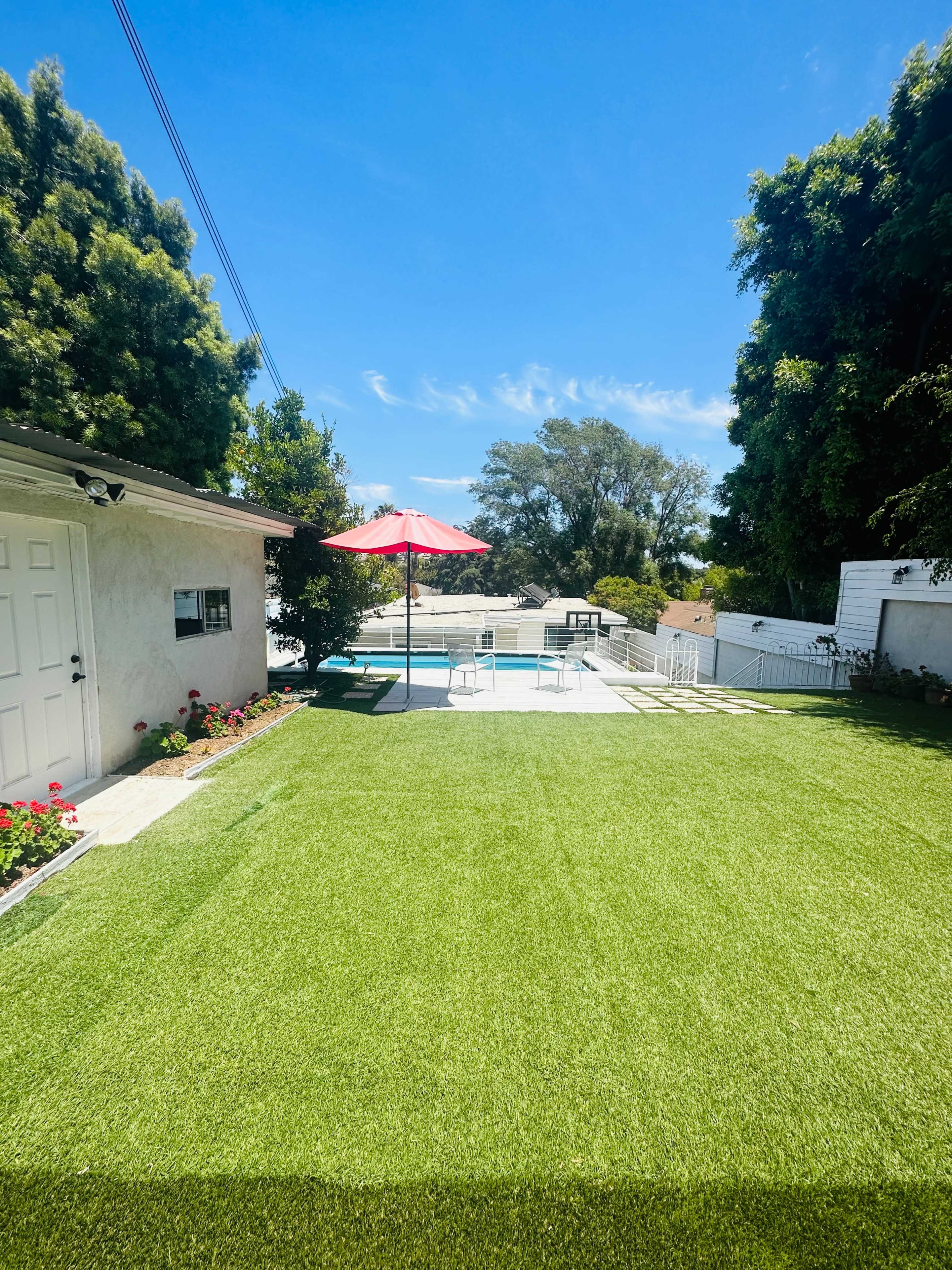 A well-maintained backyard features a swimming pool, a red umbrella, and artificial turf surrounded by greenery.
