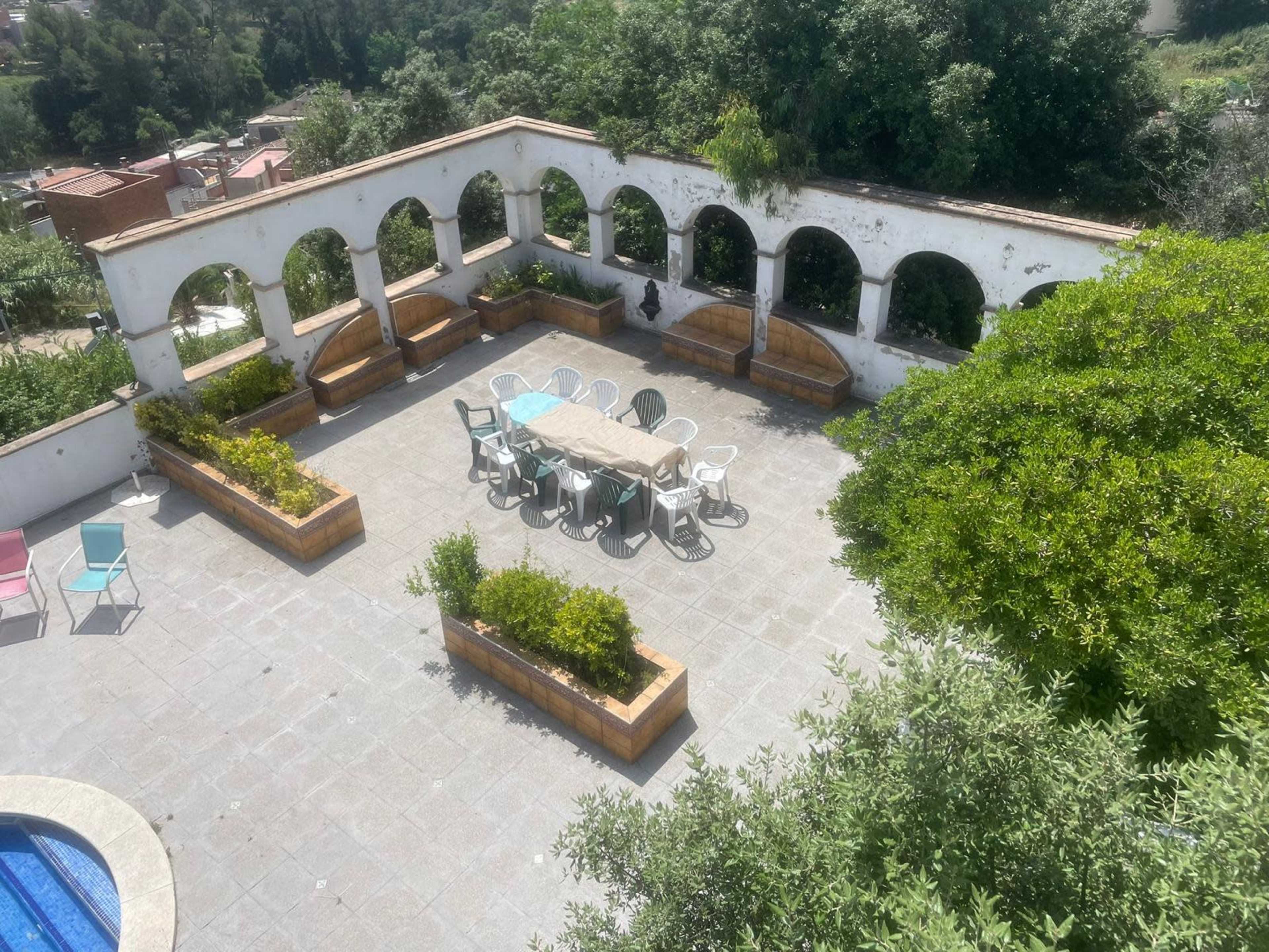 The image shows a spacious outdoor patio with arched alcoves, a large table surrounded by chairs, and trees in the background.