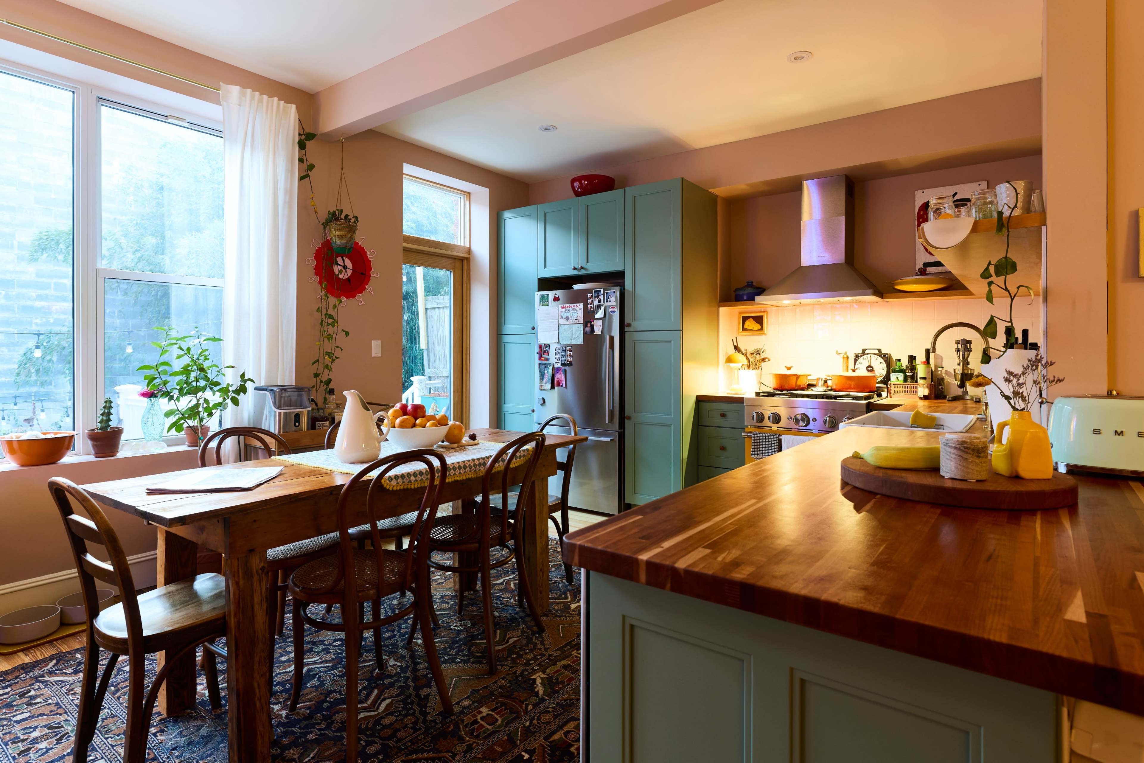 A bright kitchen features a wooden dining table surrounded by chairs, teal cabinets, and a stainless steel refrigerator, with sunlight streaming through a large window.