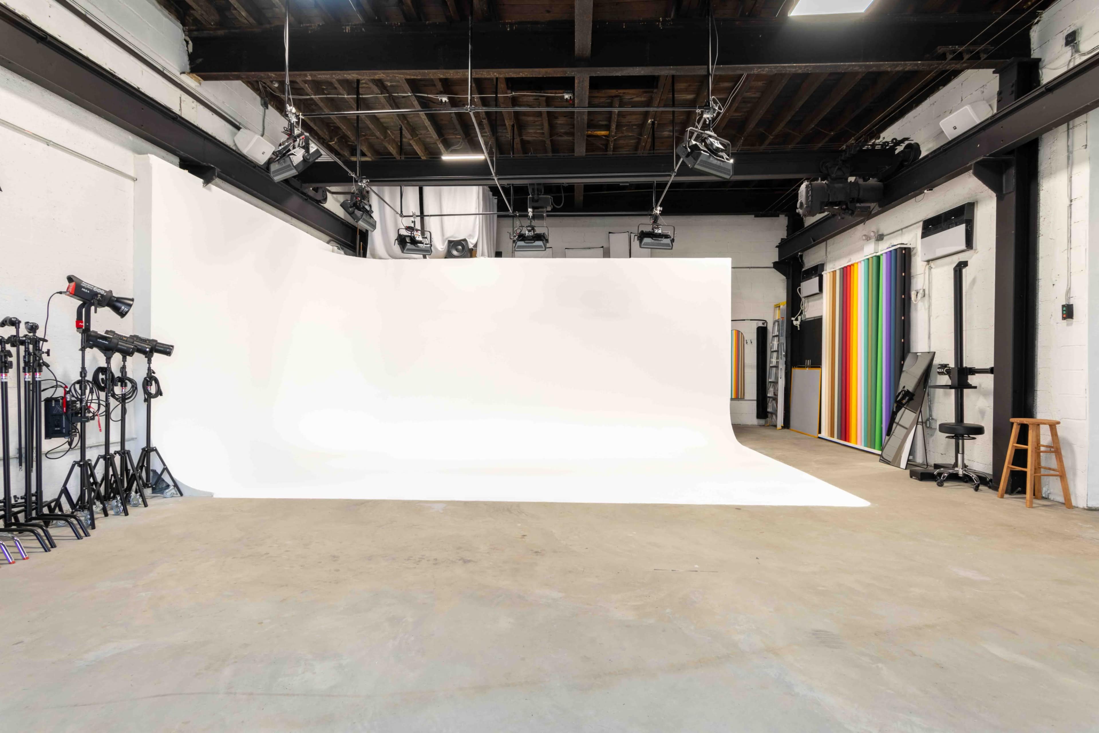 The image shows a spacious photography studio with a large white backdrop, various lighting equipment, and a colorful striped wall on one side.