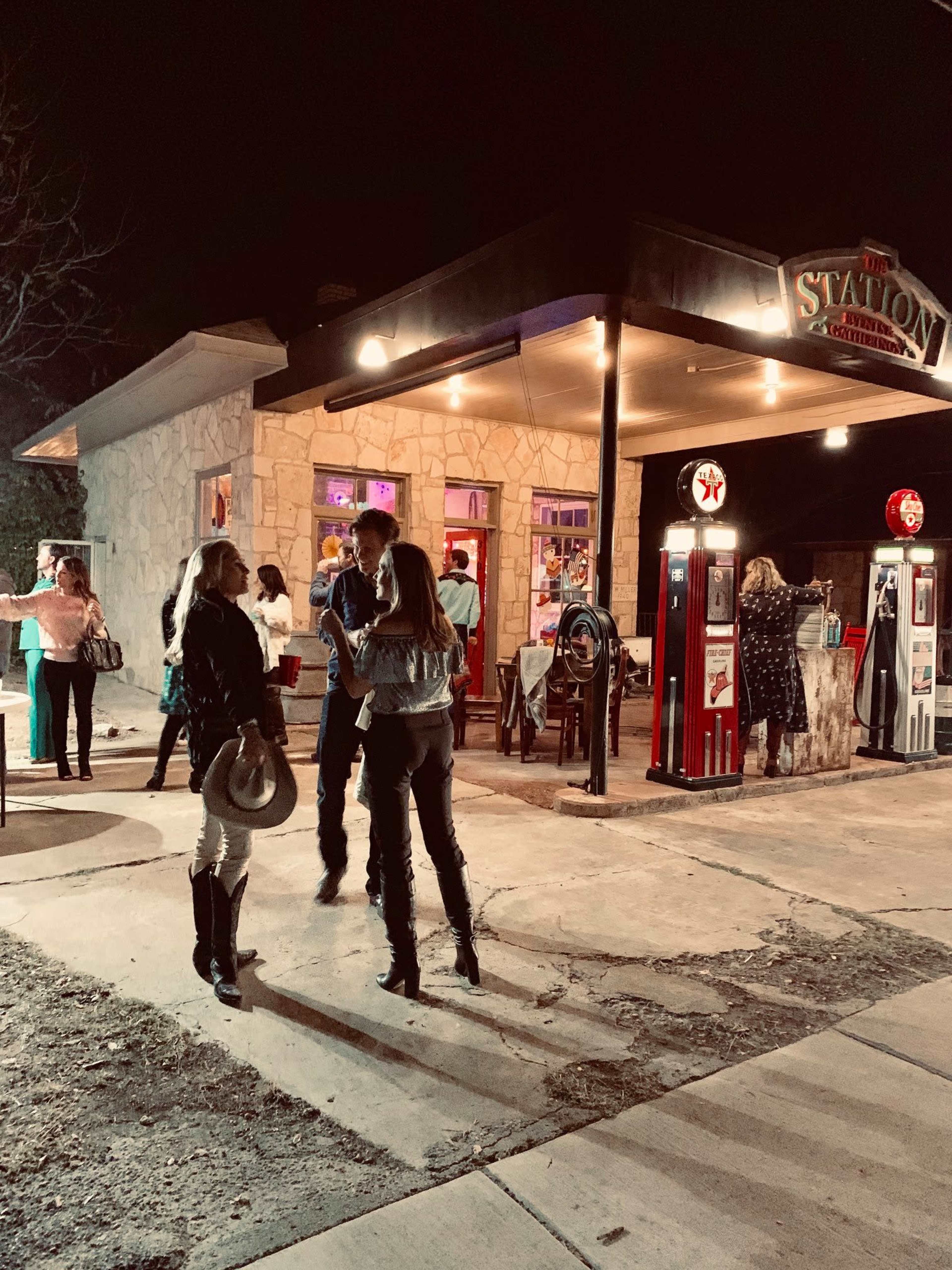 A group of people gather outside a vintage gas station at night, with colorful lights illuminating the stone building and antique gas pumps.