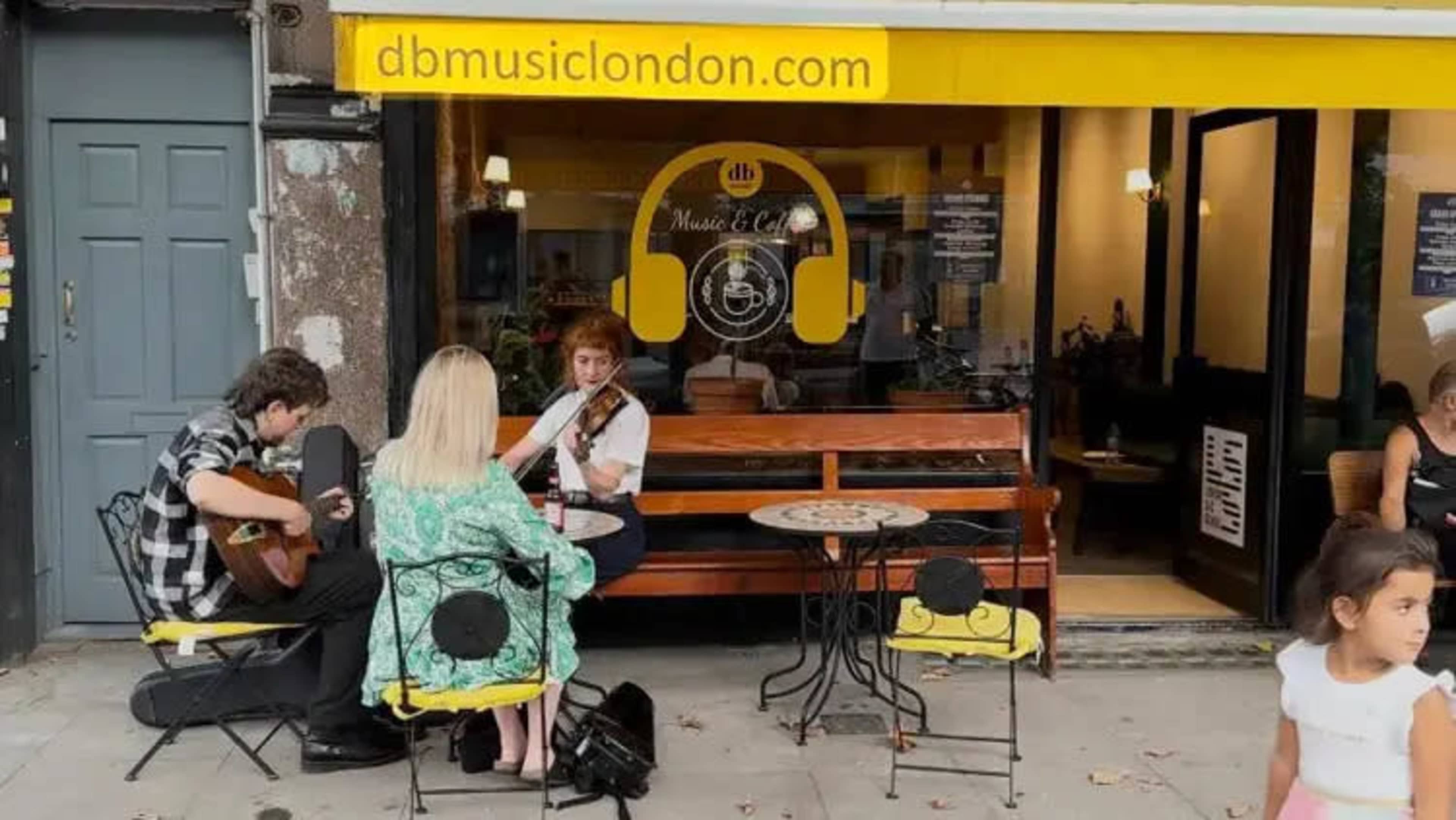 A guitarist and a violinist perform on the sidewalk outside a music café with yellow signage.