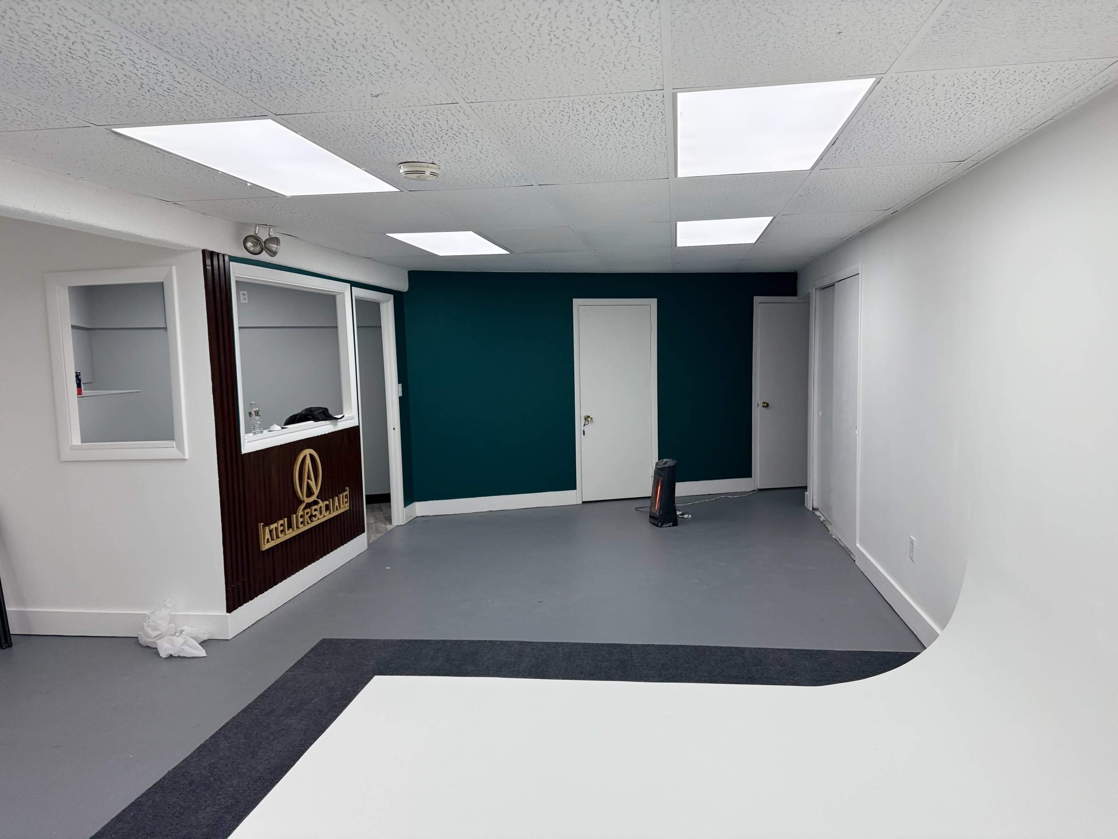 The image shows a spacious room with a teal accent wall, a white floor, and a reception area featuring a wooden counter, as well as a closed door and a vacuum cleaner in the corner.