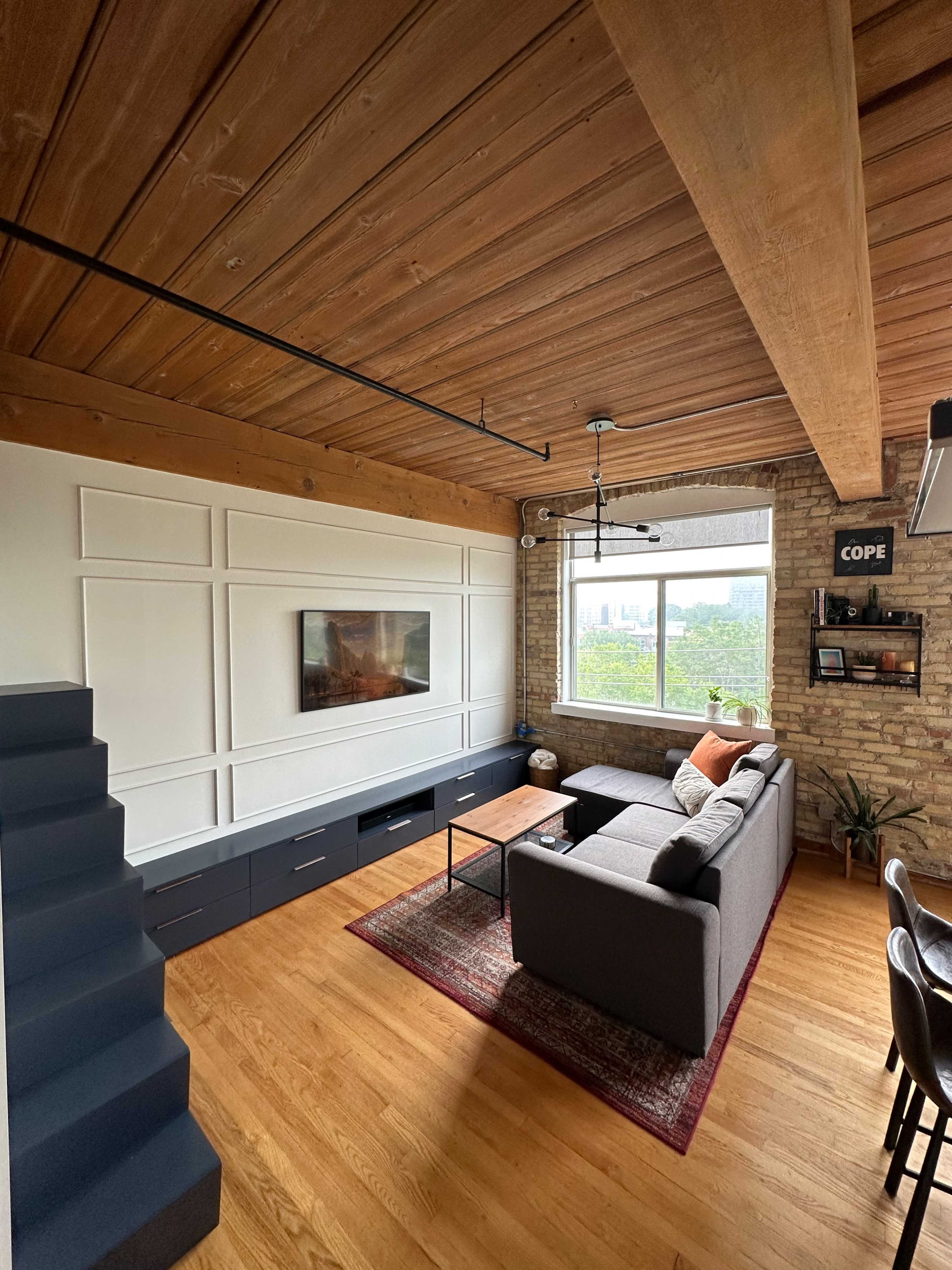 A modern living room featuring a gray sofa, a wooden coffee table, a flat-screen TV mounted on a white panel, and large windows with a view of greenery.