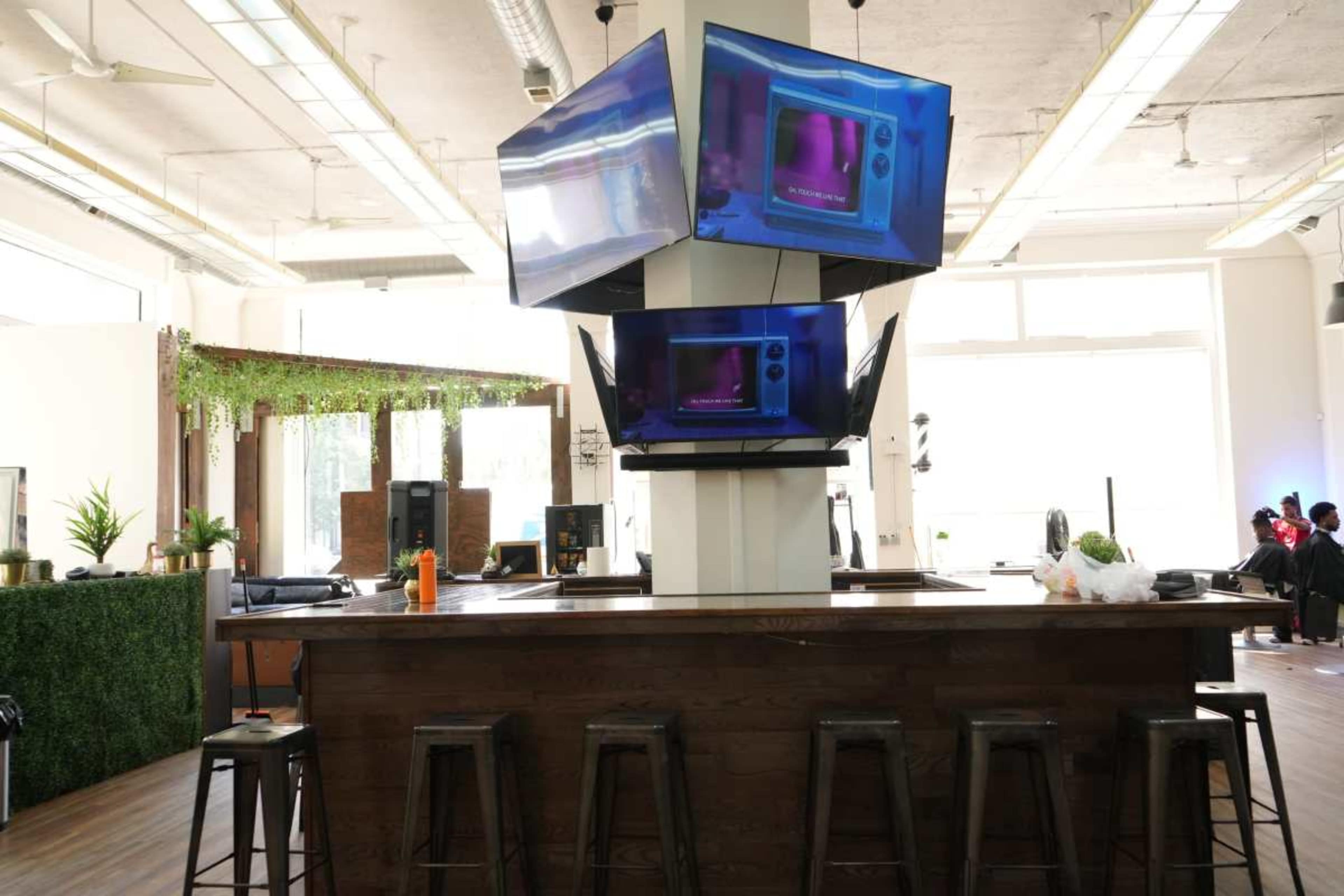 The image shows a modern bar area with multiple flat-screen TVs mounted in a geometric arrangement above a wooden counter, surrounded by indoor plants and bright natural light.