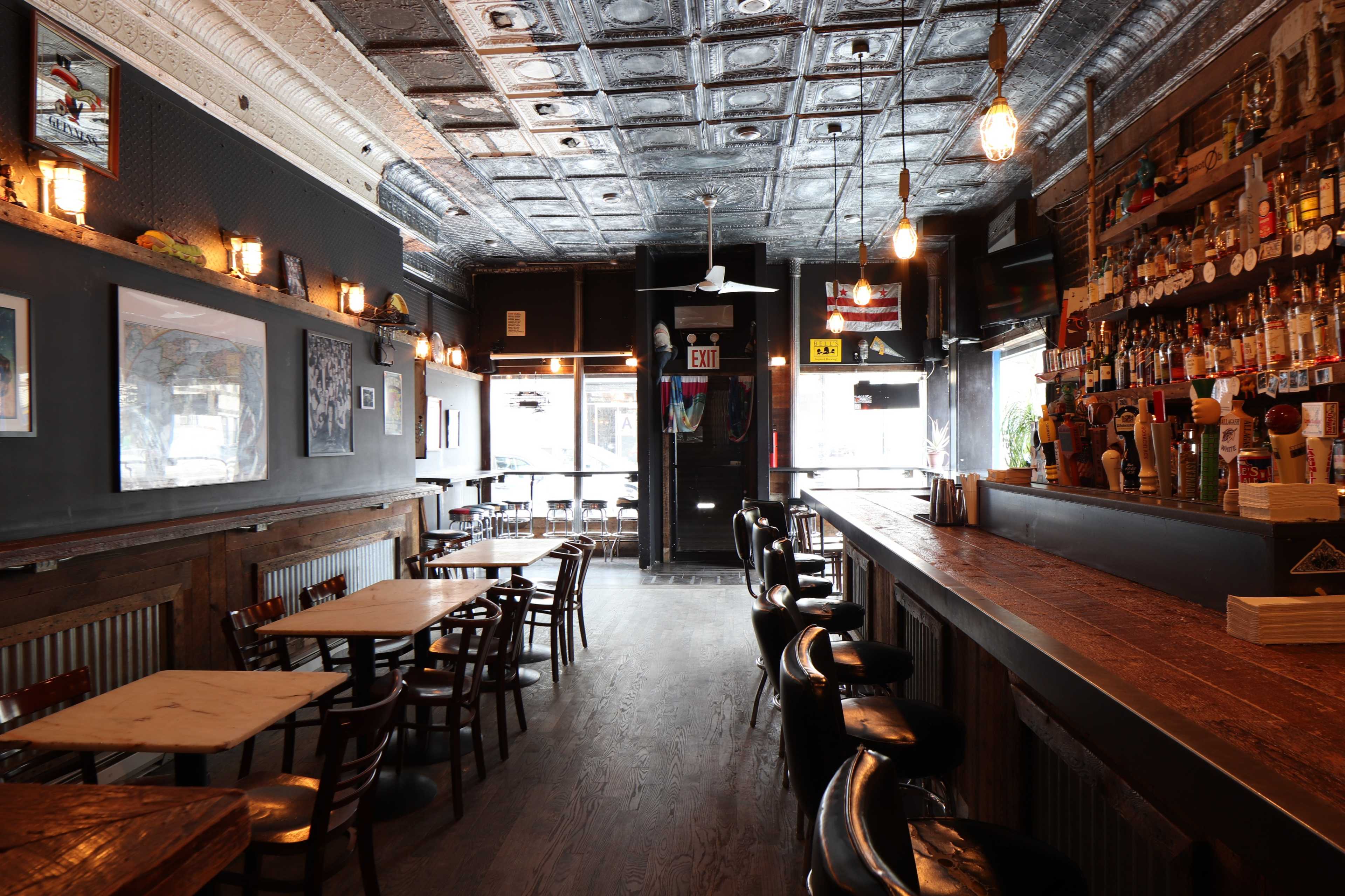 The interior of a bar with wooden tables and chairs, a long wooden bar counter, and decorative ceiling tiles.