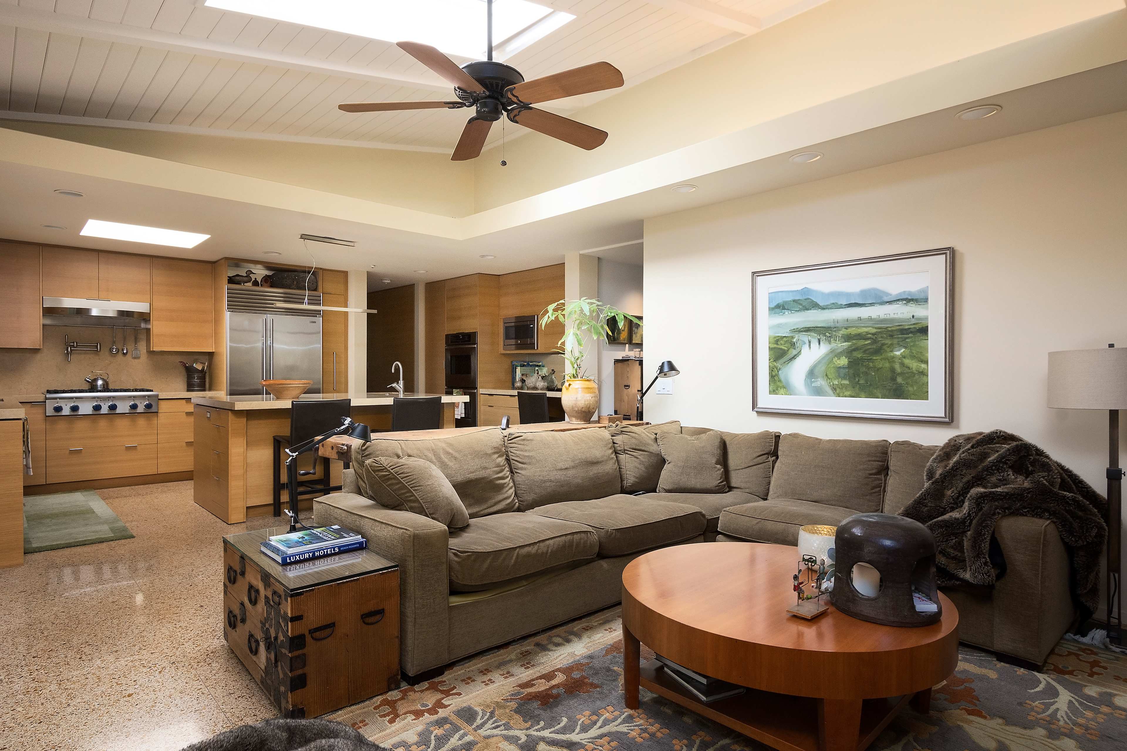 A living room with a large brown sectional sofa, a round wooden coffee table, and a view into a modern kitchen with wooden cabinets and stainless steel appliances.
