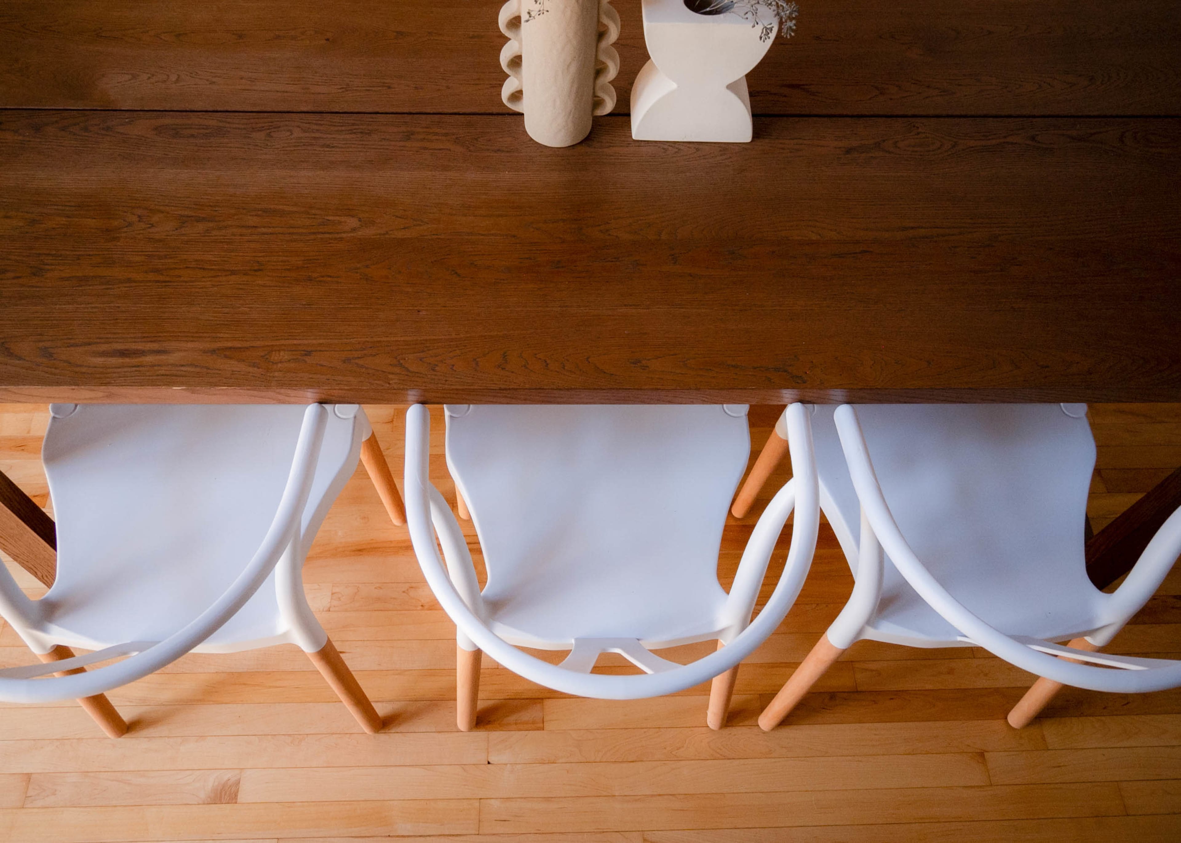 Three white chairs are positioned under a wooden table on a light wooden floor, with a vase decoration visible on the table's surface.