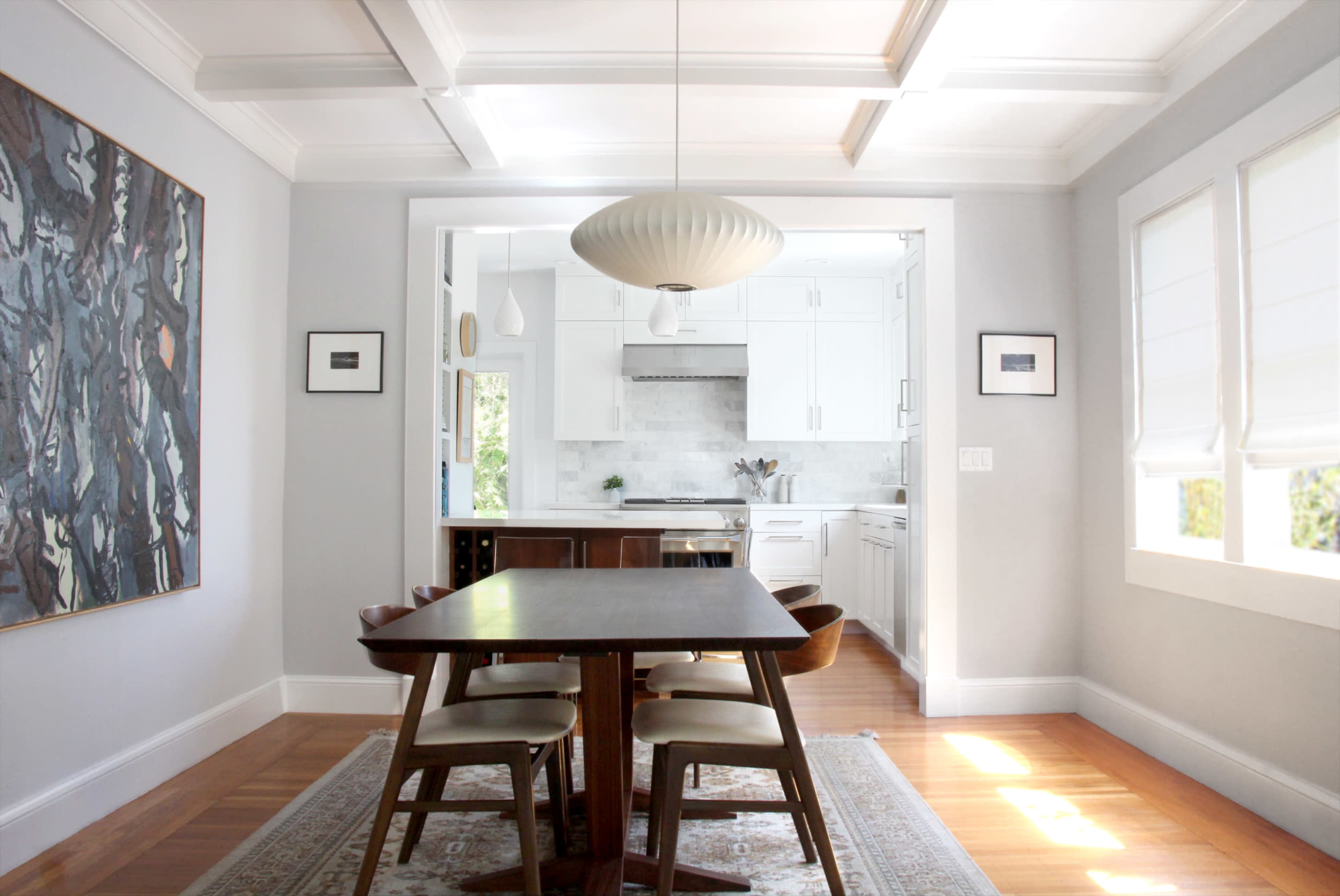 A dining area features a wooden table surrounded by chairs, with a modern light fixture hanging above and a view into a white kitchen through an open doorway.