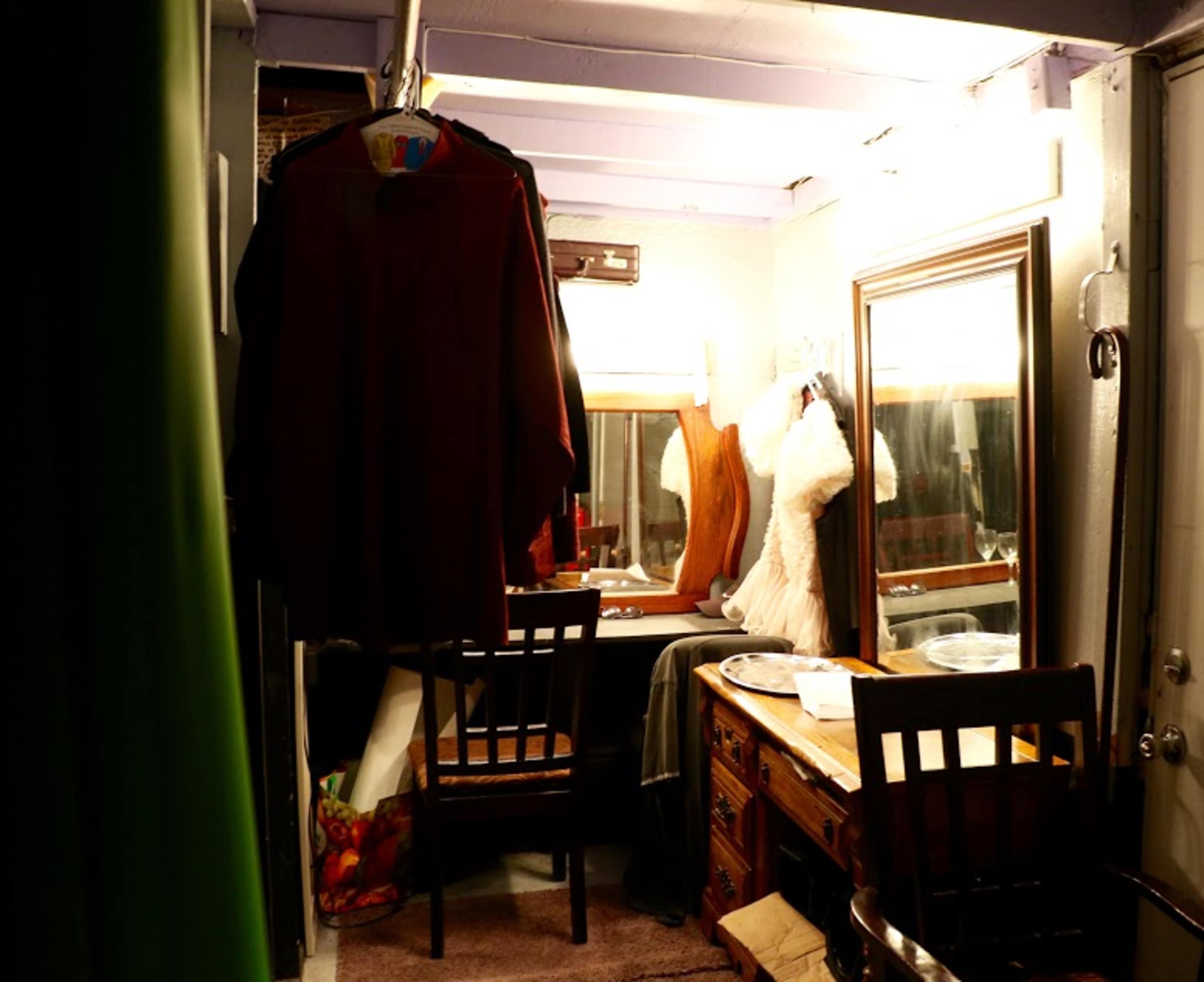 The scene depicts a dimly lit dressing room with clothes hanging on a rack, a vanity mirror, and a wooden table surrounded by chairs.