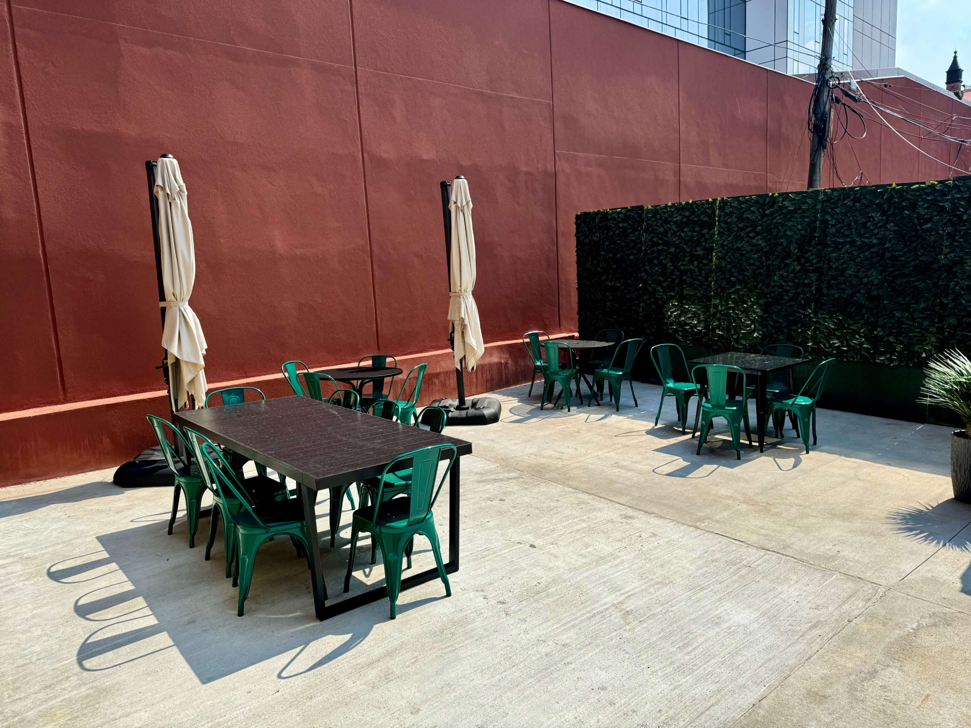 An outdoor dining area with several black tables and green chairs surrounded by a red wall and a tall hedge.