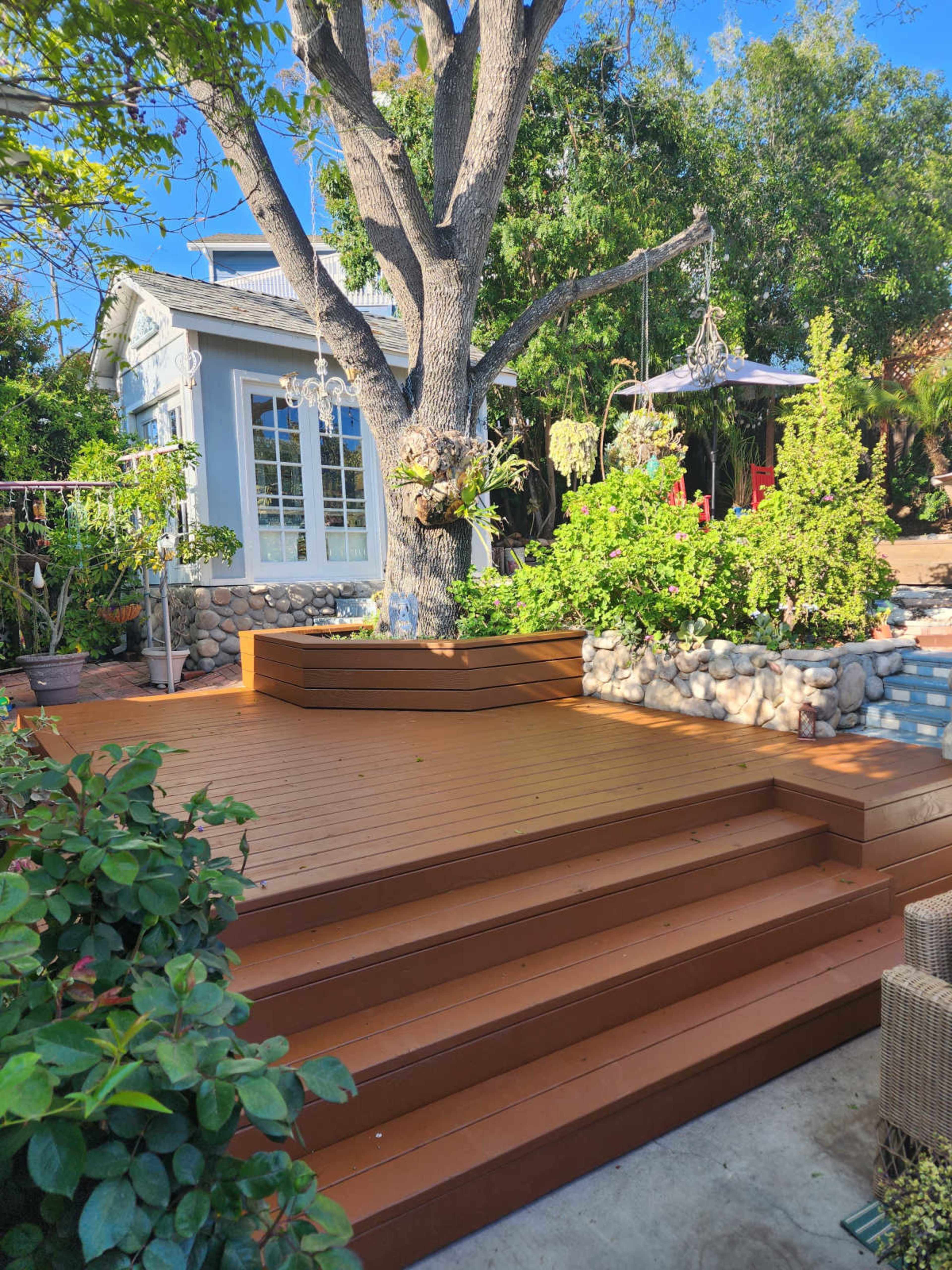 A spacious wooden deck features steps leading up to a large tree surrounded by various plants and a decorative stone border.