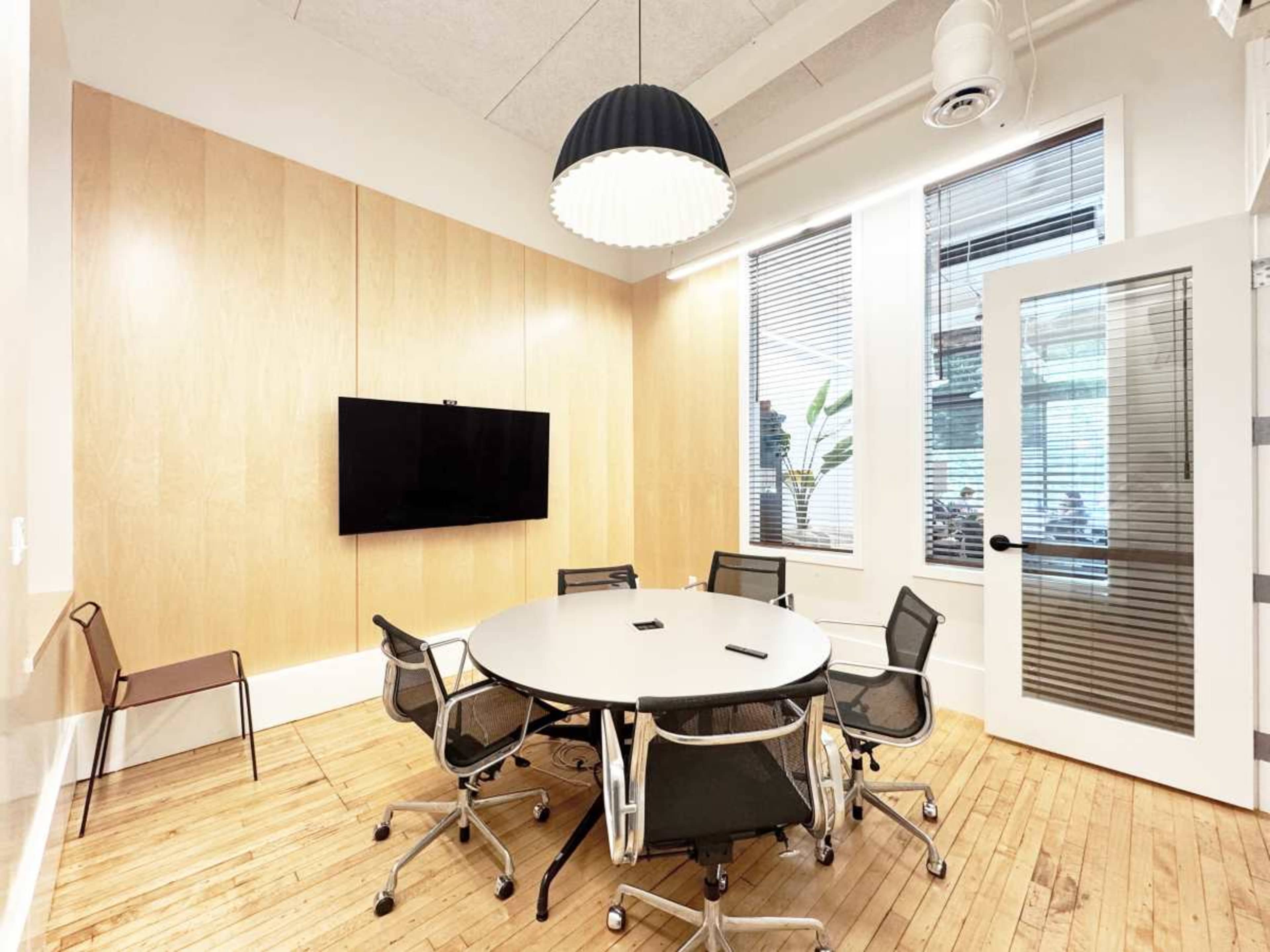 A small conference room features a circular table surrounded by six chairs, a wall-mounted TV, and large windows with blinds.