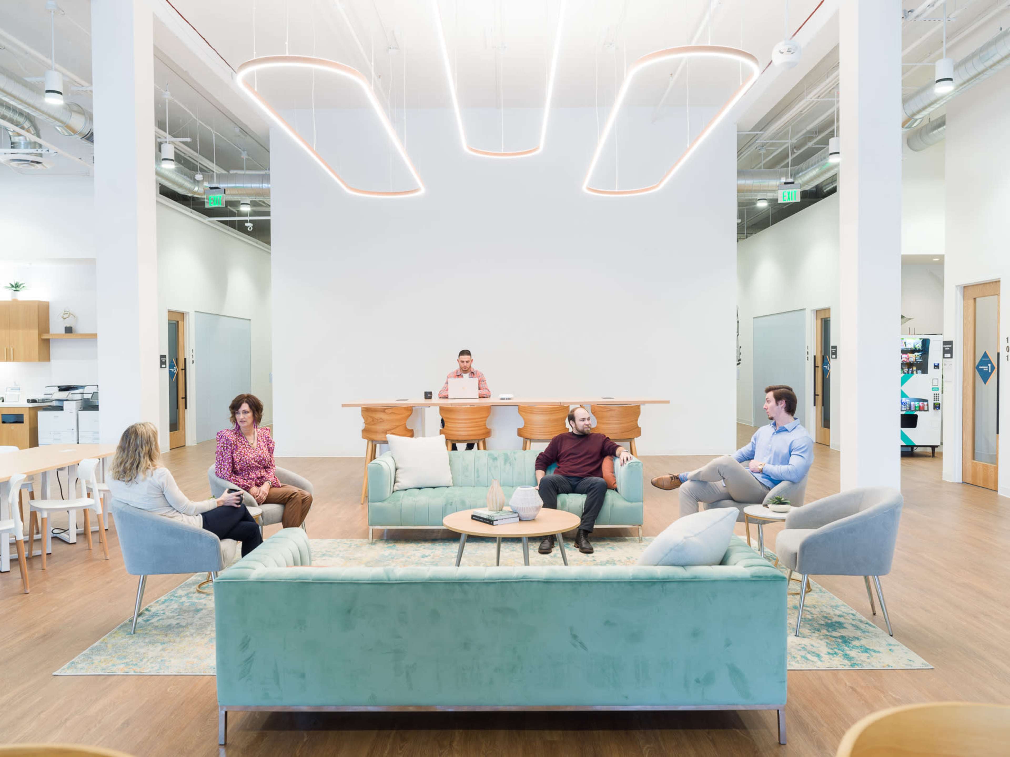 The image shows a modern office space with a seating area where five people are engaged in conversation while one person stands at a reception desk in the background.