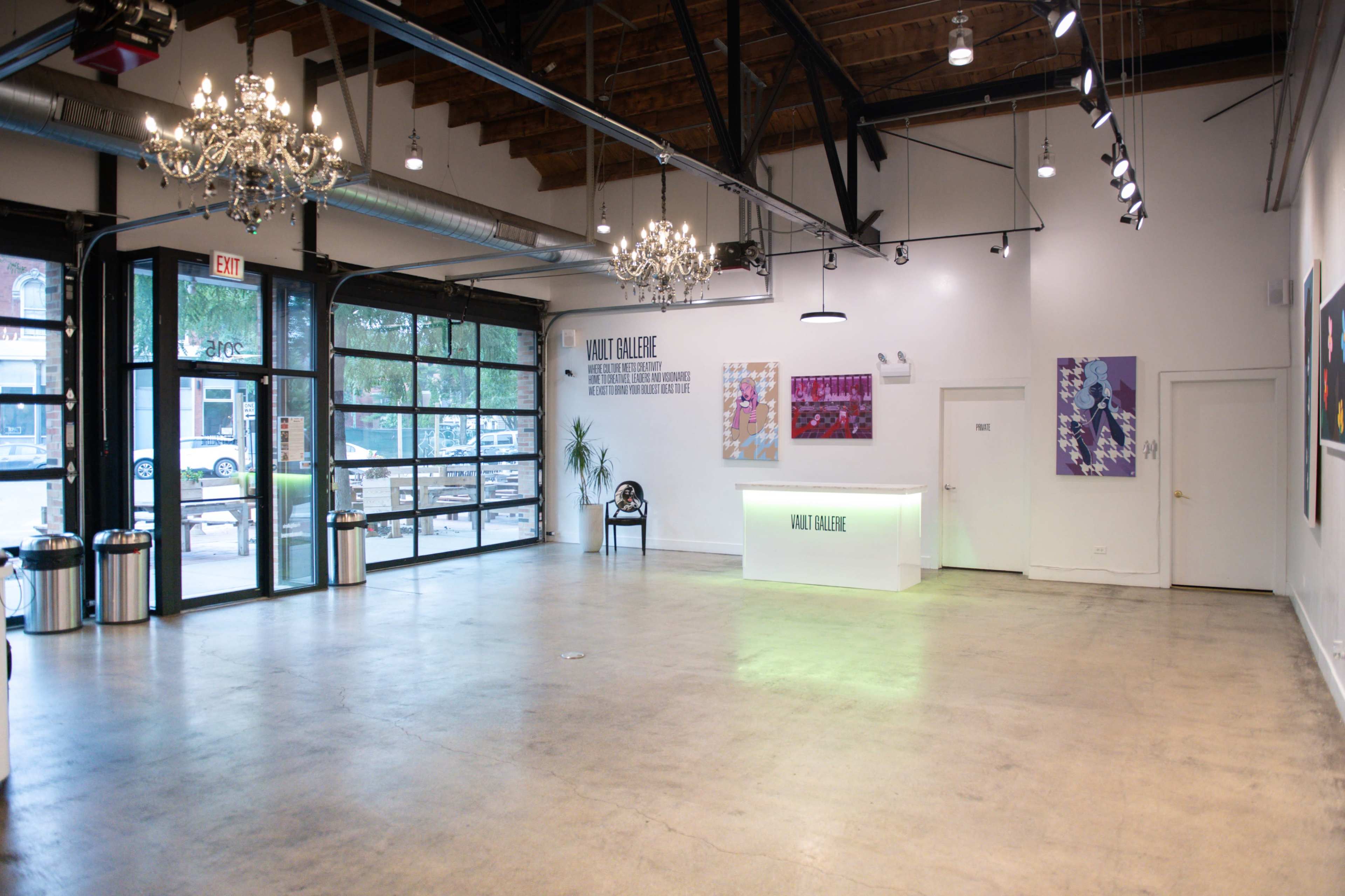 The image shows a spacious, modern gallery with a reception area, large windows, and artwork displayed on the walls.