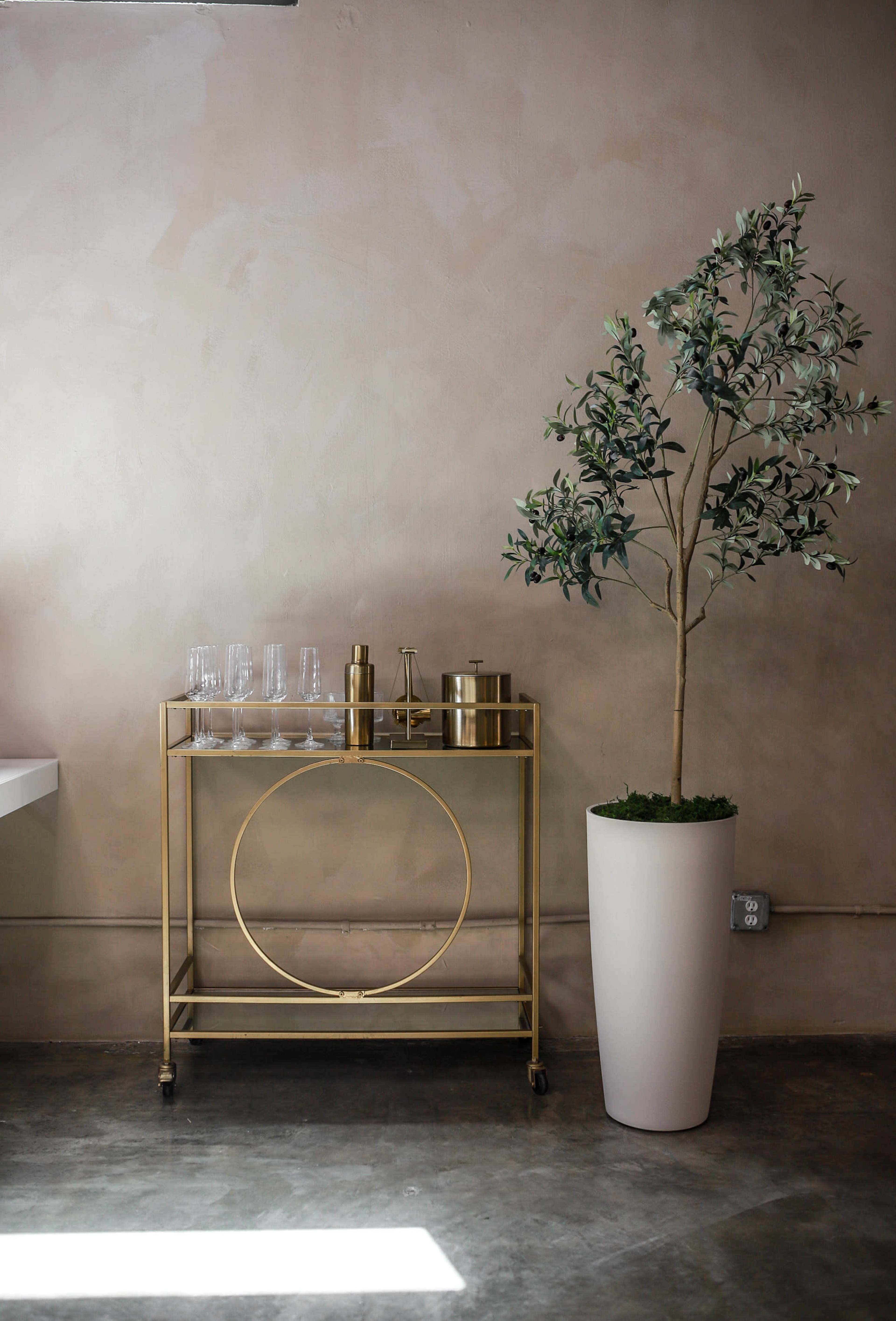 A gold bar cart with glassware and a cocktail set is positioned next to a large white planter containing a faux tree against a beige wall.