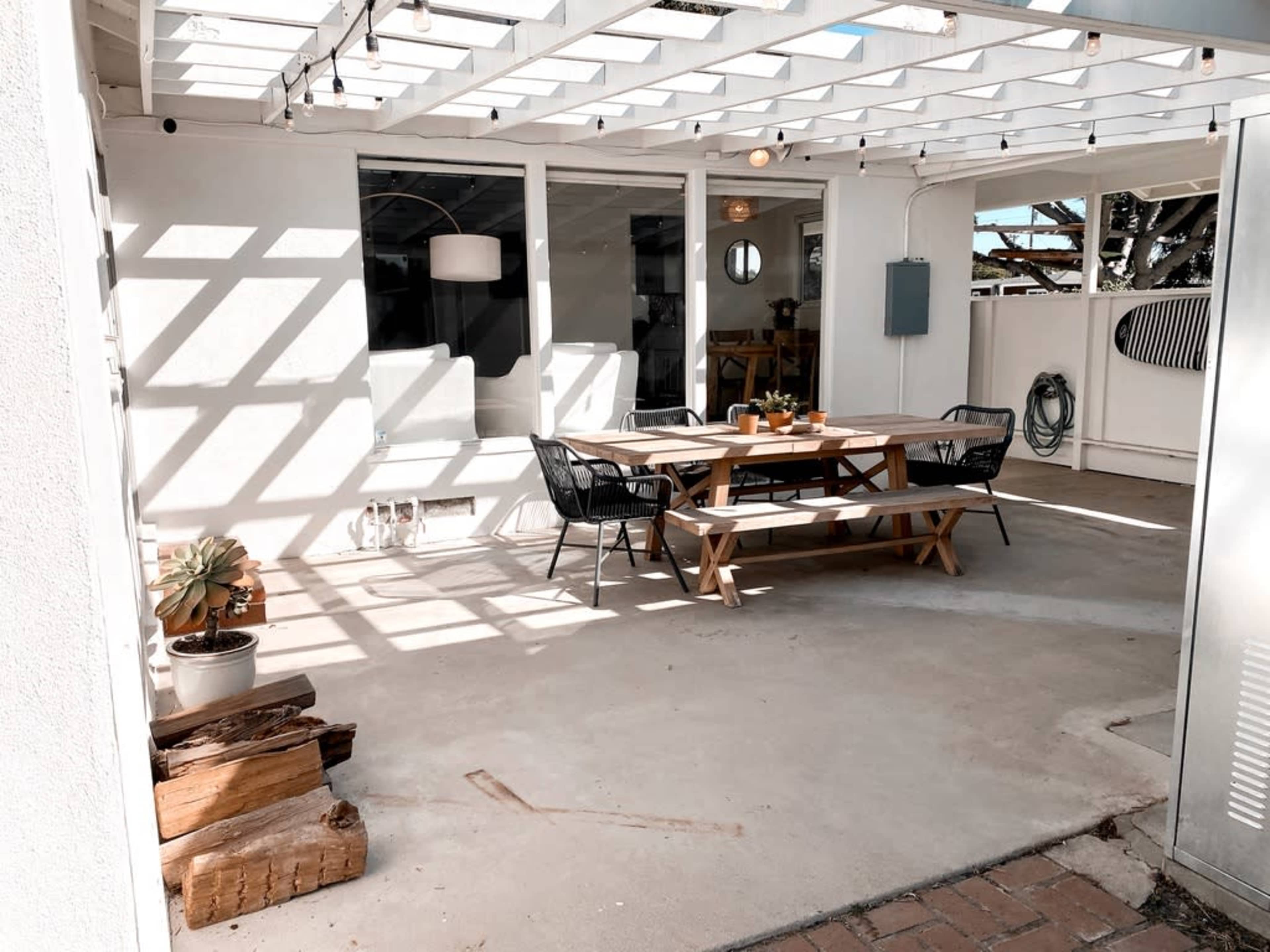 A patio area features a wooden table with benches, surrounded by potted plants and sunlight casting shadows through a slatted overhead structure.