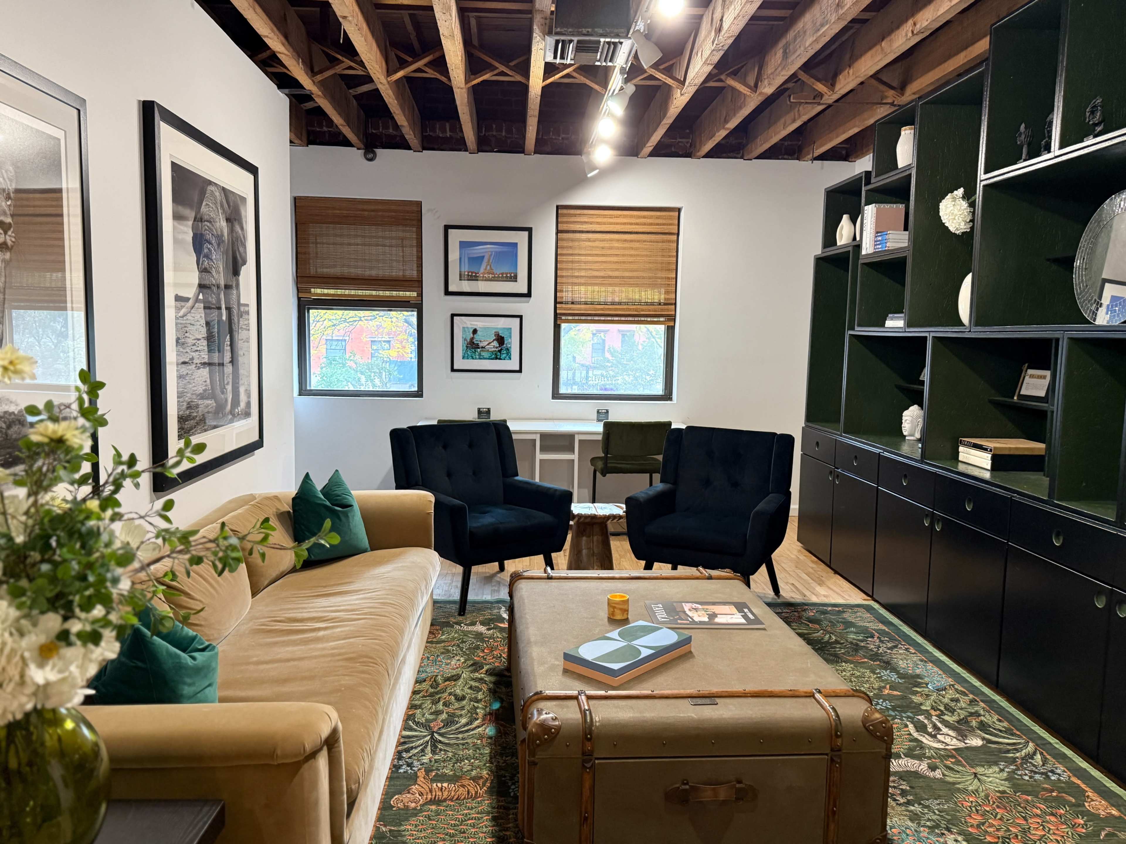 The image shows a modern living space with a tan sofa, two dark armchairs, a green bookshelf, and decorative elements, all under a wooden ceiling.