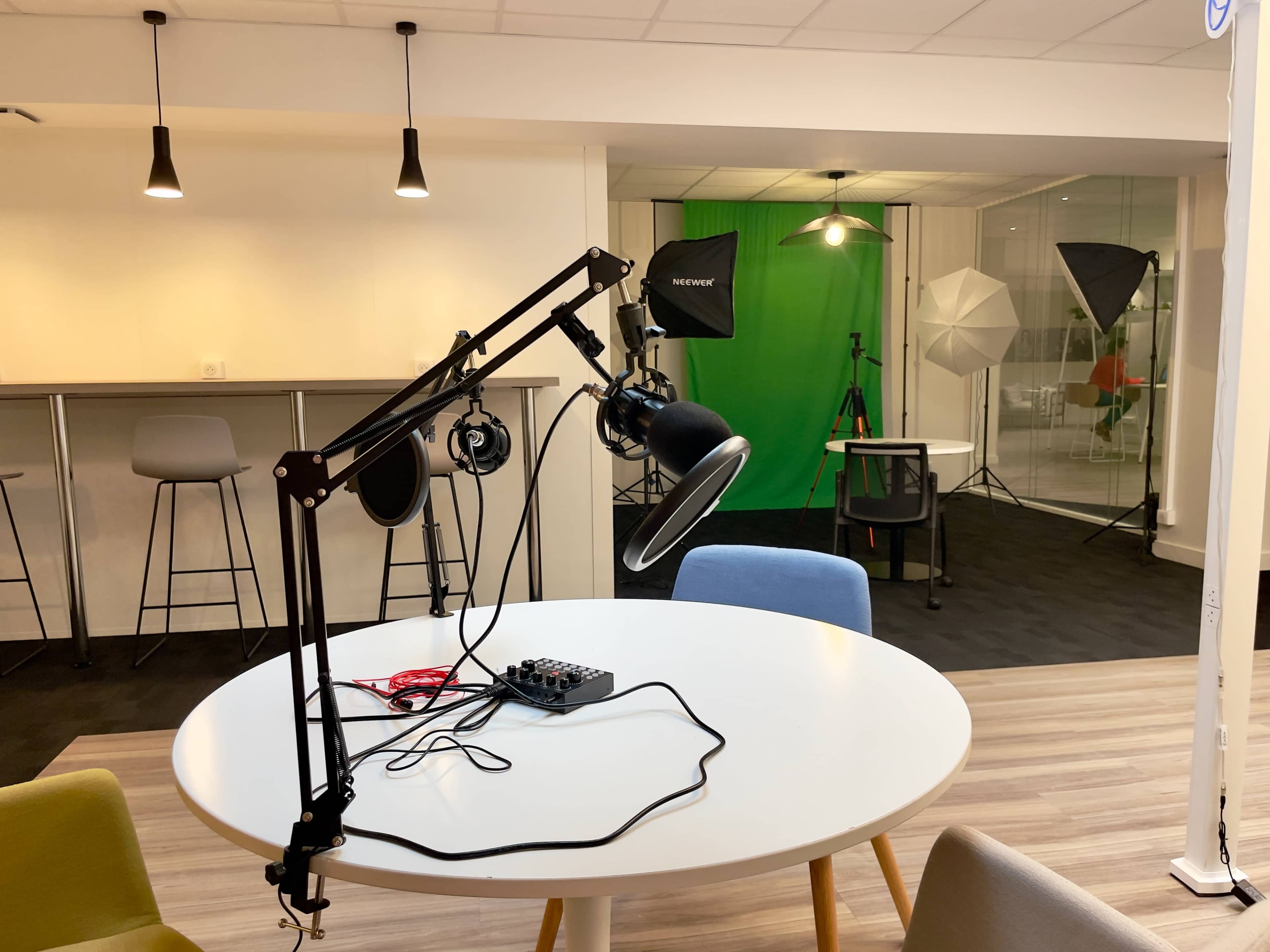 The image shows a studio setup with a round white table in the foreground, featuring a microphone and headphones, and a green screen along with lighting equipment in the background.