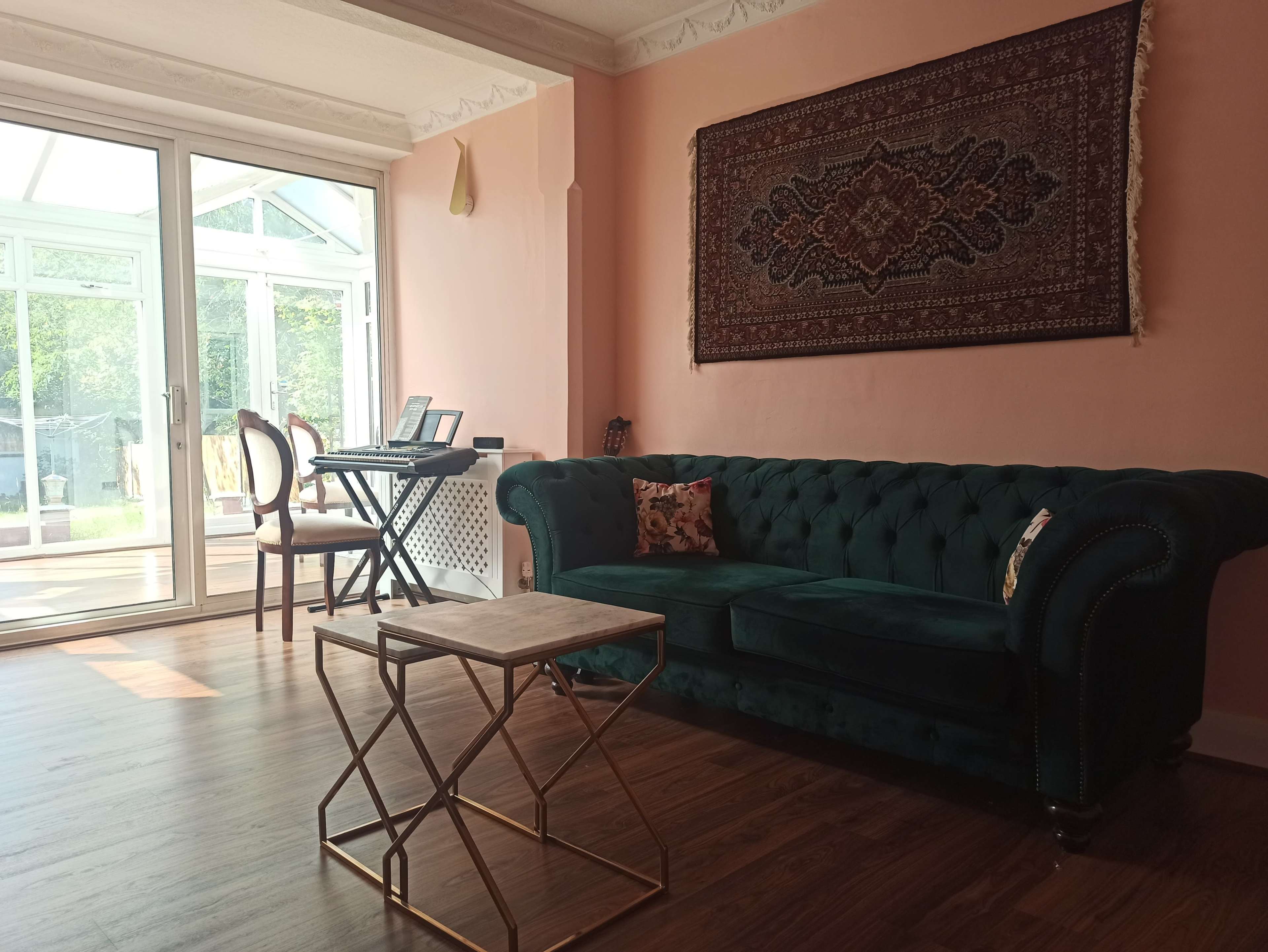 A spacious room with a green tufted sofa, a small gold coffee table, and a glass door leading to a sunlit conservatory.