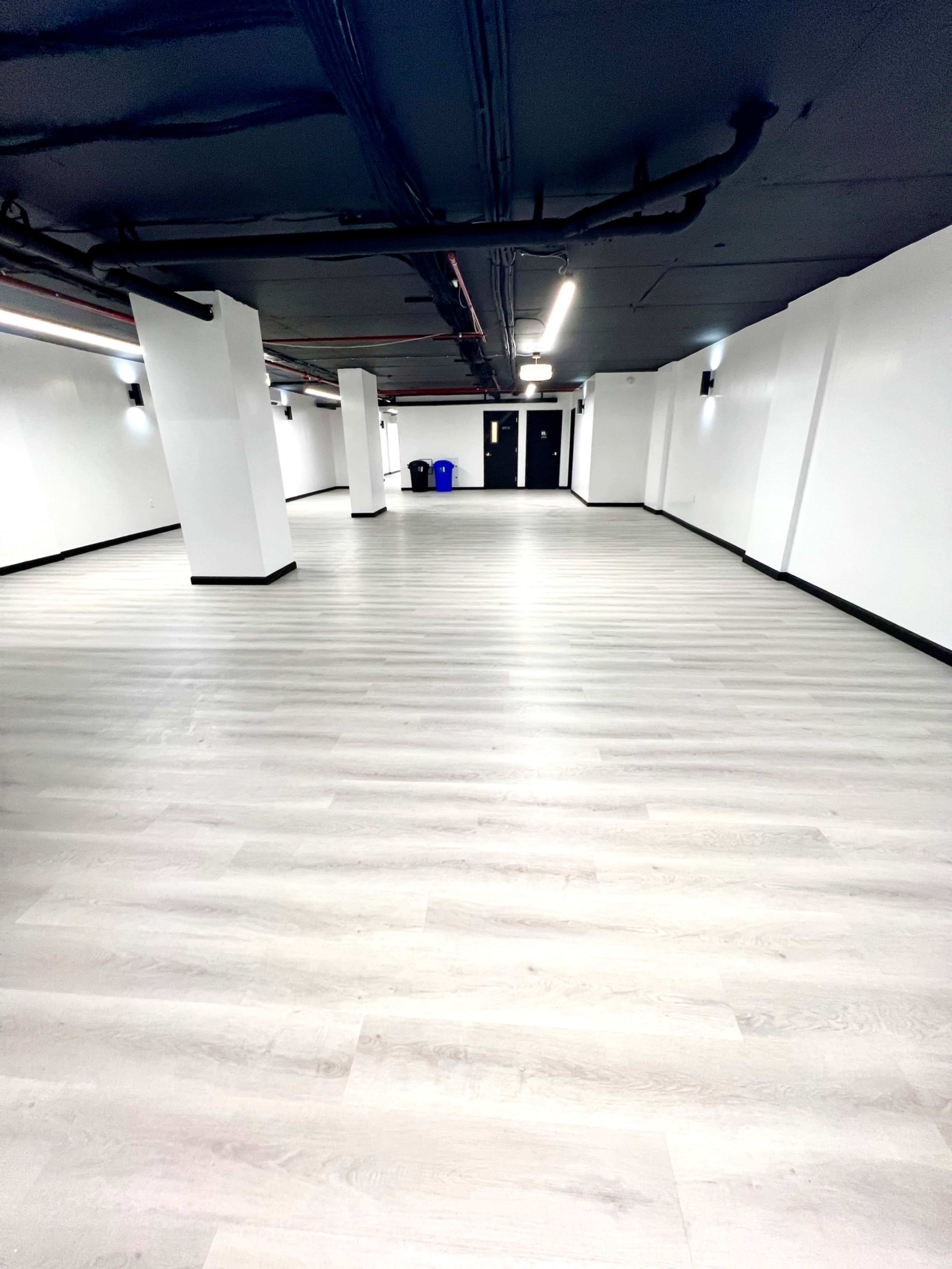 A large, empty room with light-colored wooden flooring and white walls, featuring black accents and minimal lighting fixtures.