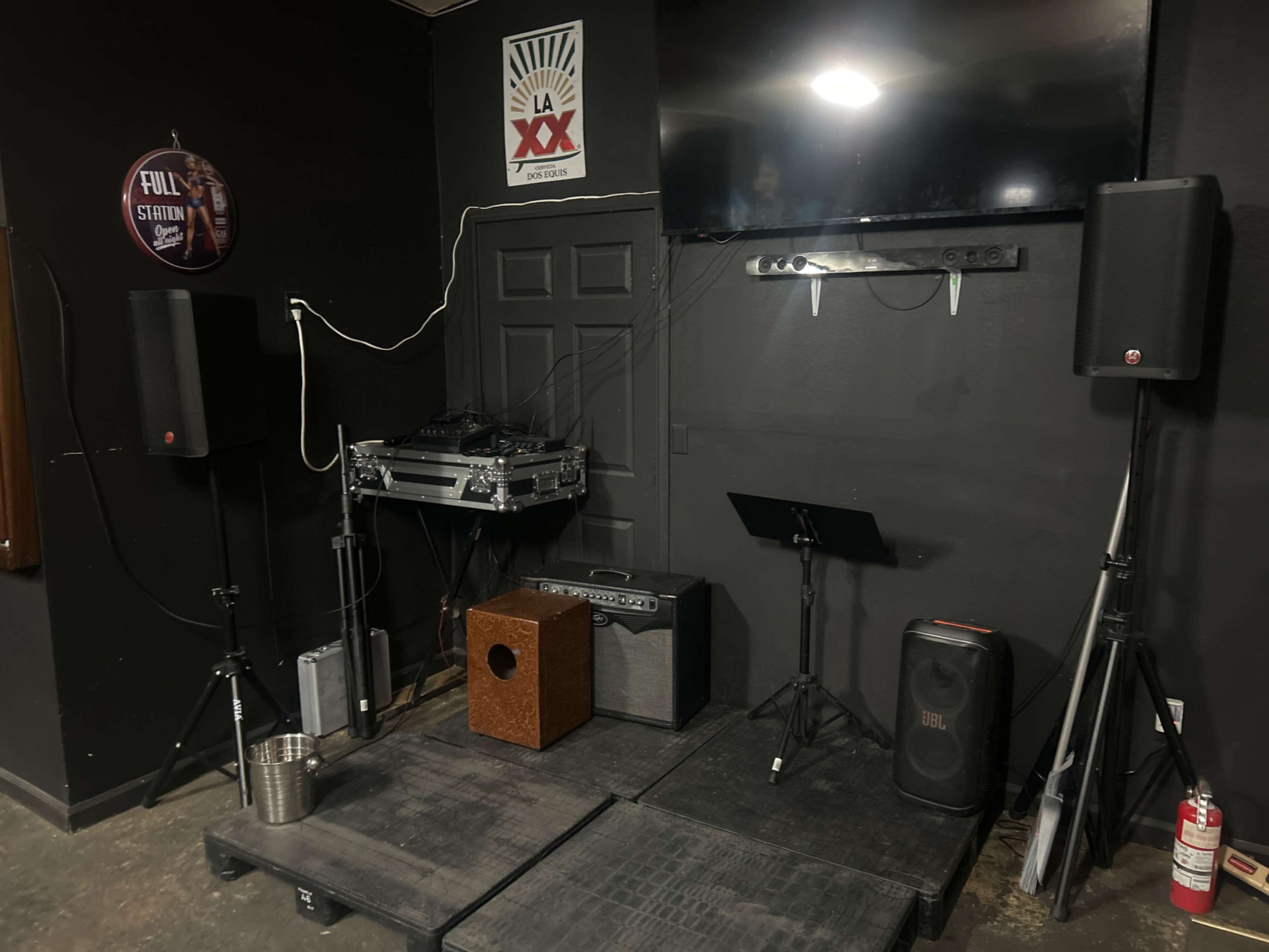 The image shows a small performance area with speakers, a DJ setup, a microphone stand, and a television mounted on the wall.