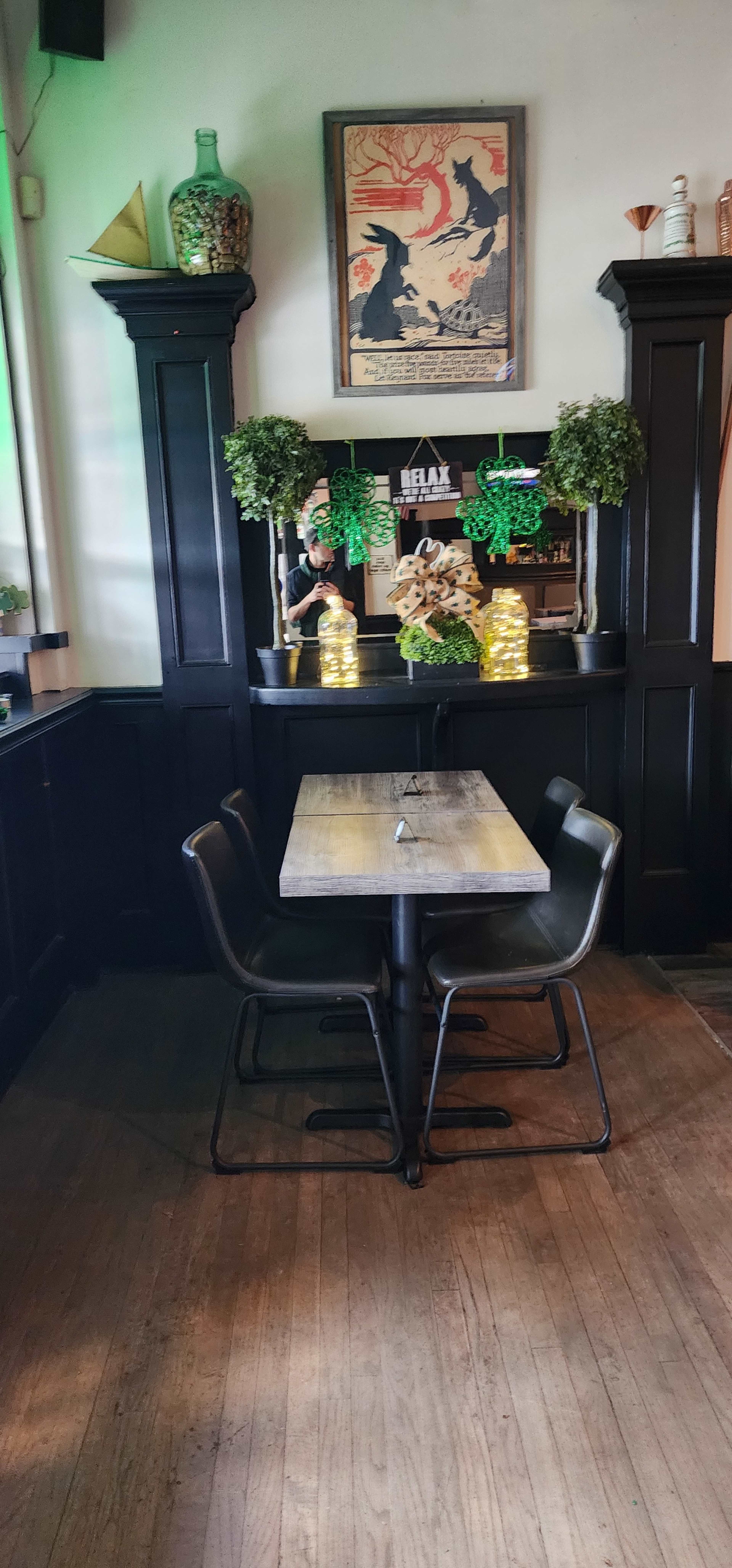 A small wooden table with black chairs is positioned in a dimly lit corner of a cafe, decorated with potted plants and lights.